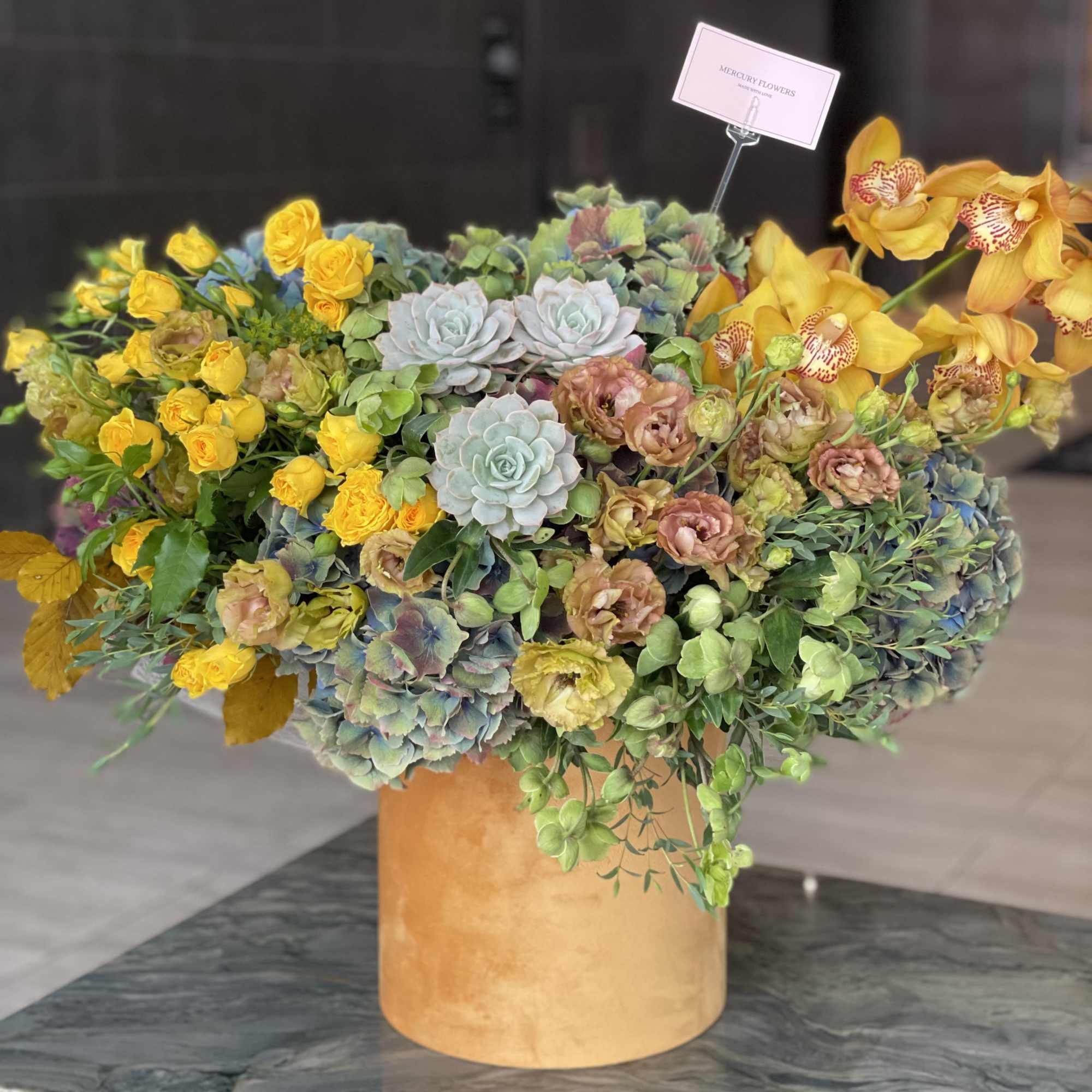 Large mixed floral arrangement in a gold vase with yellow orchids and succulents