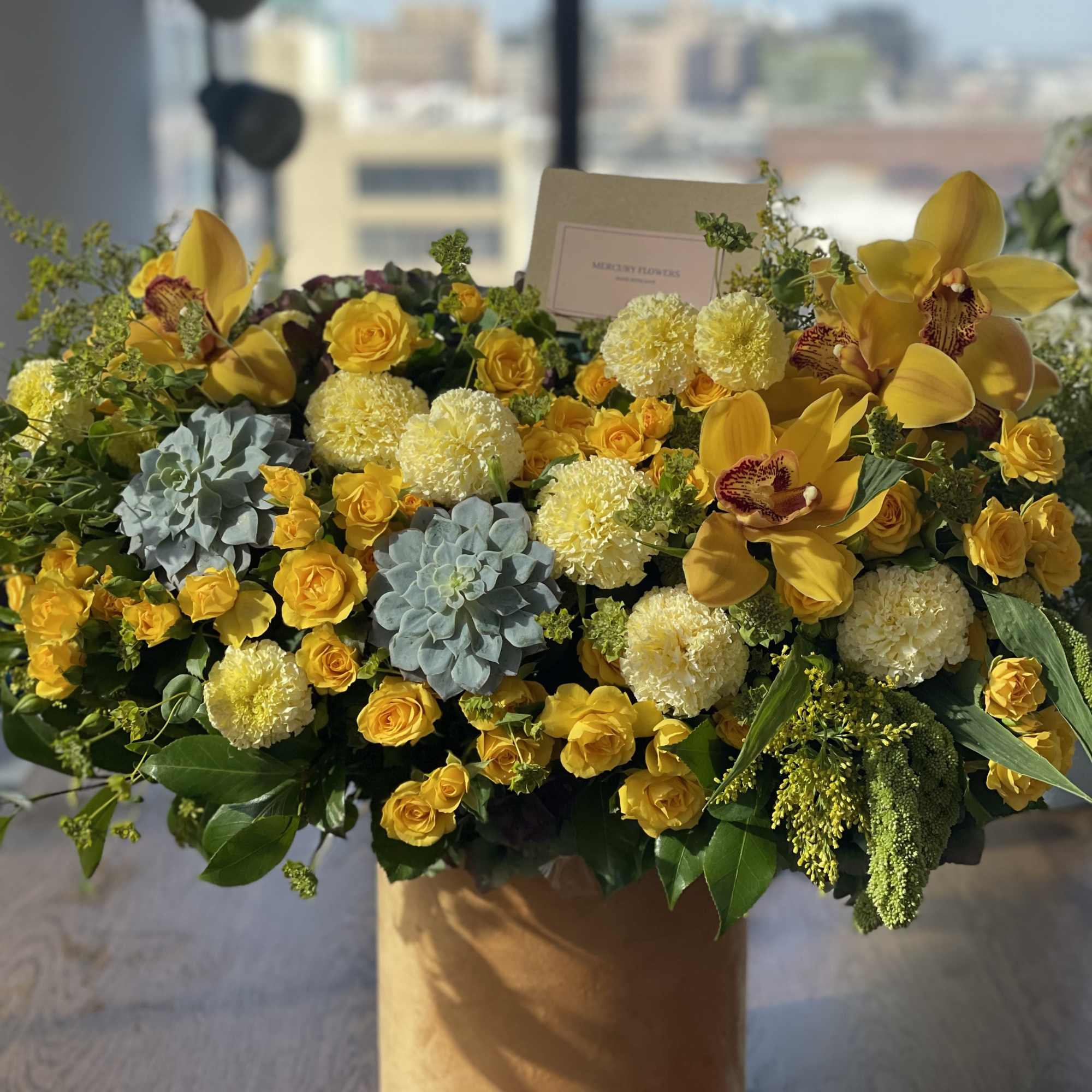 Large yellow floral arrangement in a tan box with roses and orchids