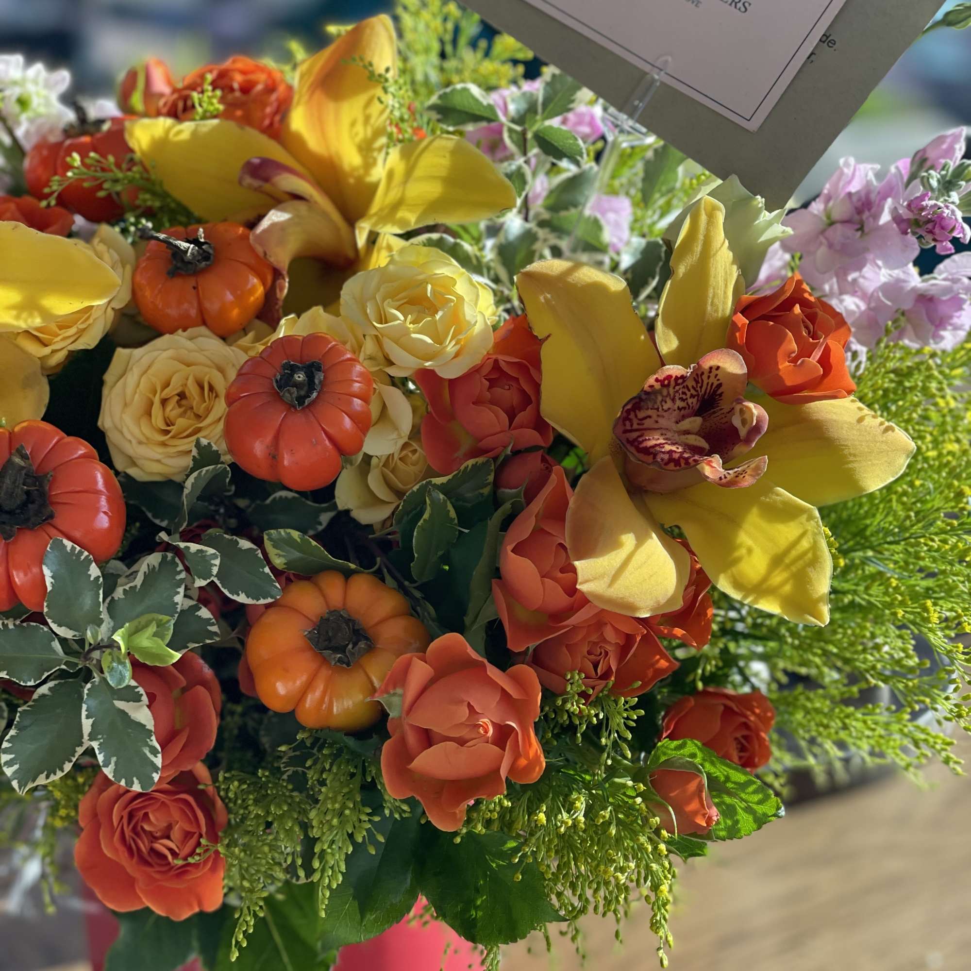Bright mixed bouquet with yellow lilies, orange roses, and small pumpkins in a pink vase.