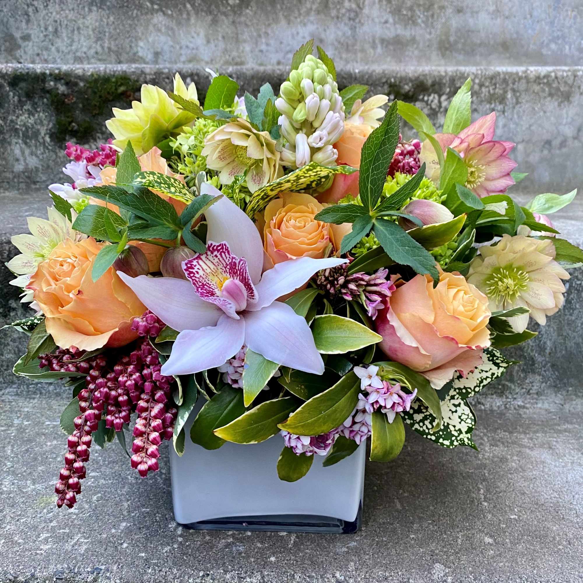 Mixed floral arrangement in a white square vase with peach roses and a lavender orchid