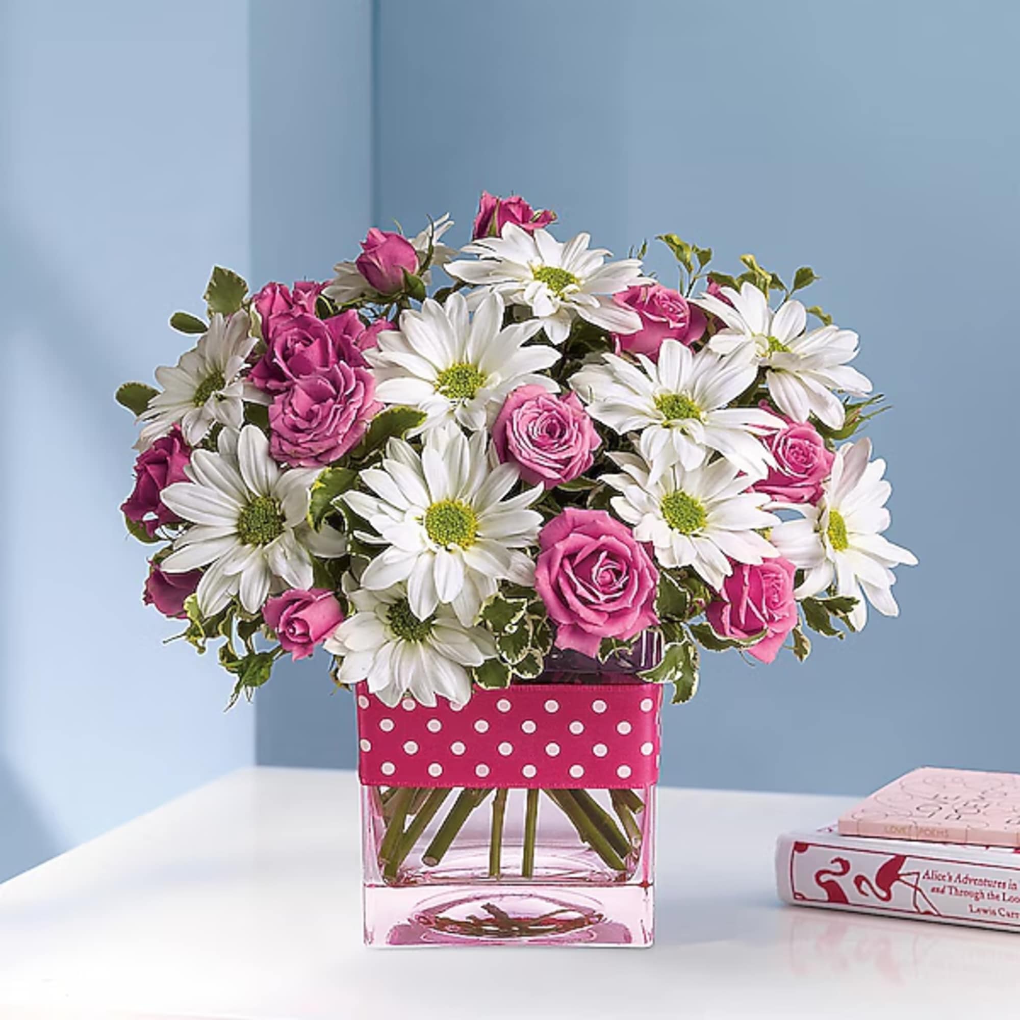Pink roses and white daisies in a clear square vase with a polka-dot ribbon