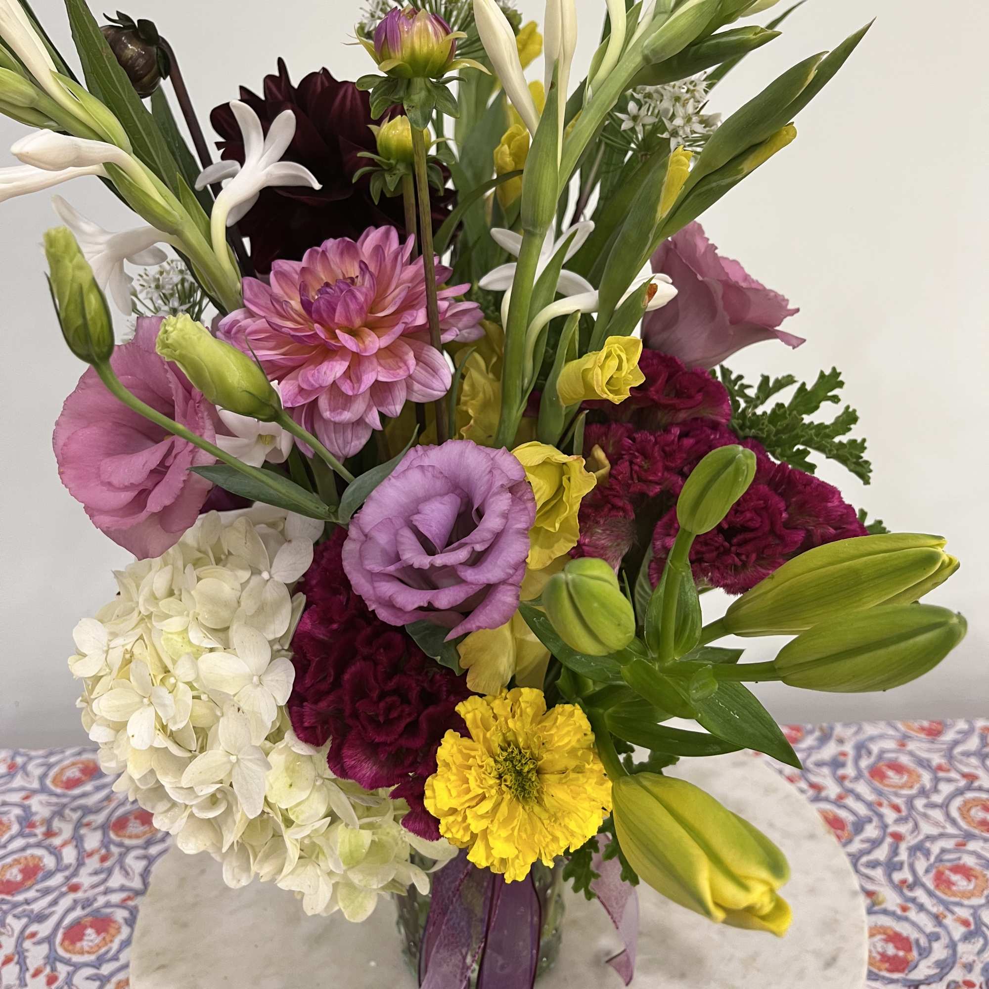 Tall mixed arrangement with purple lisianthus, white hydrangea, yellow marigolds, lilies and pink dahlia in a glass vase.