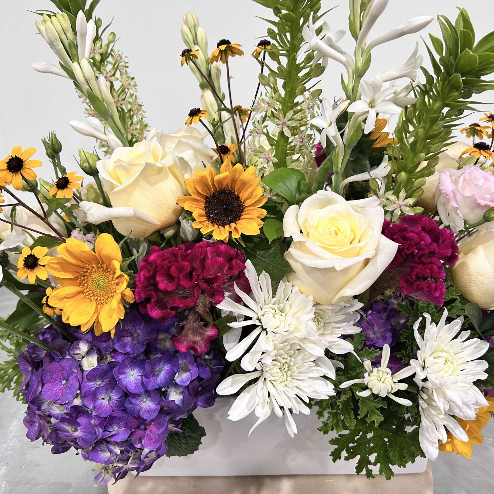 Mixed bouquet with white roses, purple hydrangeas, and yellow daisy-like flowers in a white container.