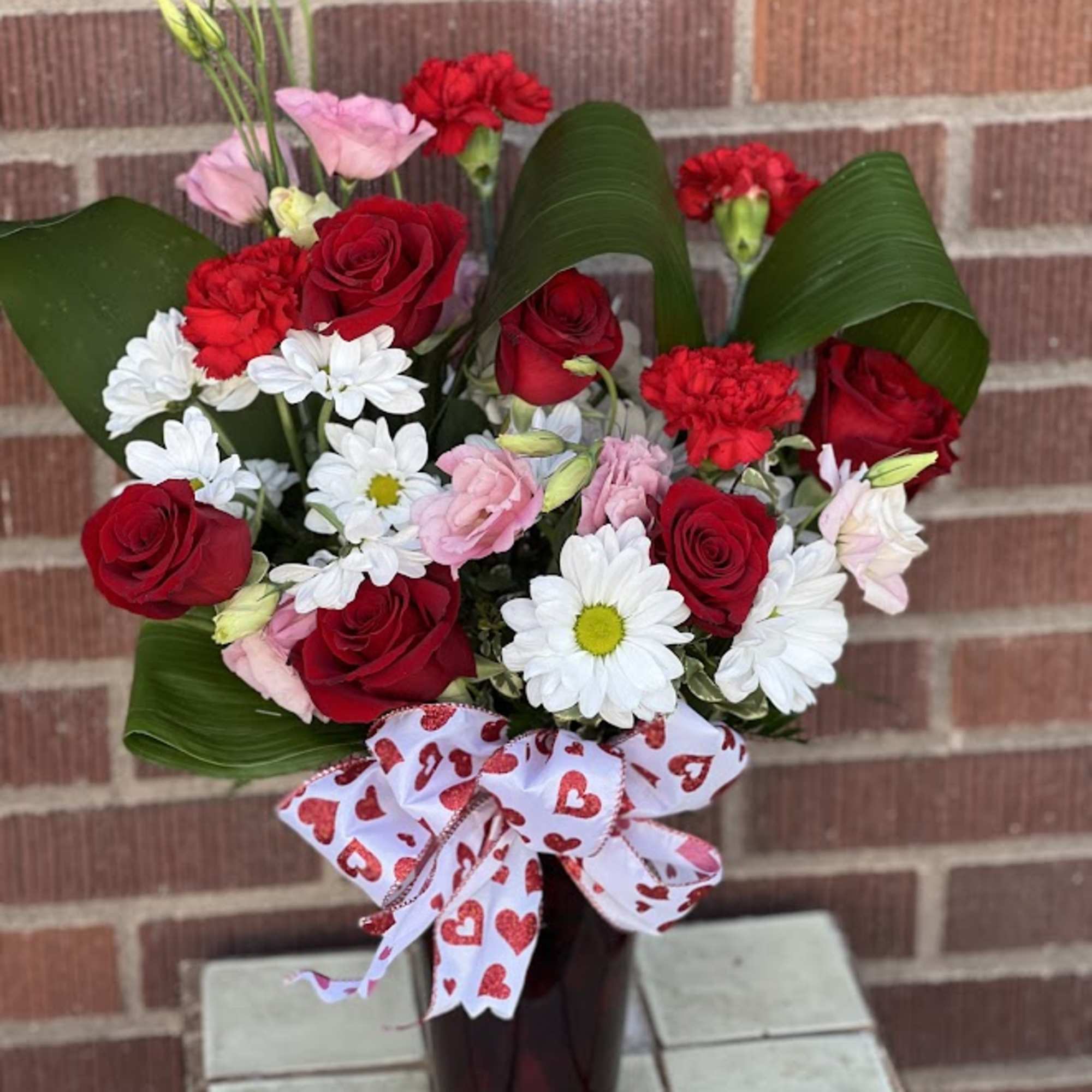 Red roses, pink blooms, and white daisies in a dark vase with a heart ribbon