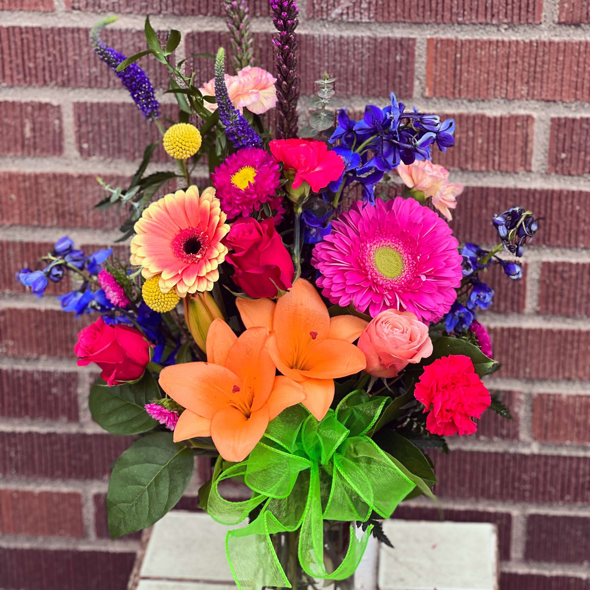 Bright mixed bouquet with lilies, roses, gerbera daisies, and a green ribbon in a glass vase