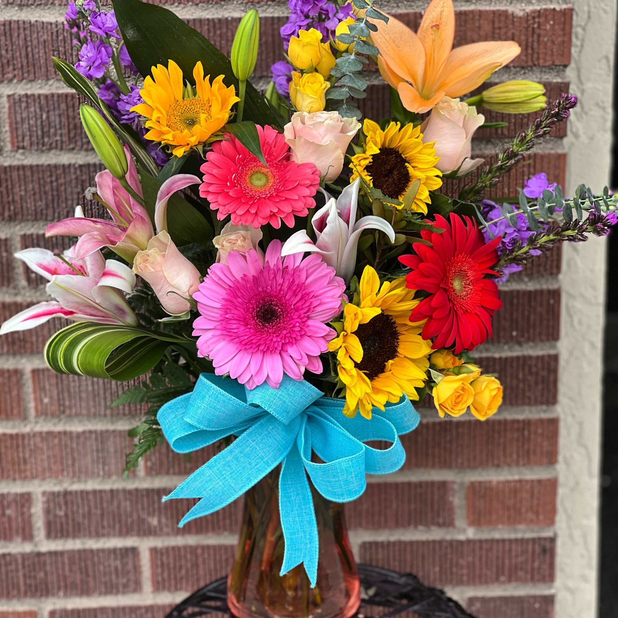 Colorful mixed bouquet with gerbera daisies, lilies, roses, and a blue ribbon in a glass vase