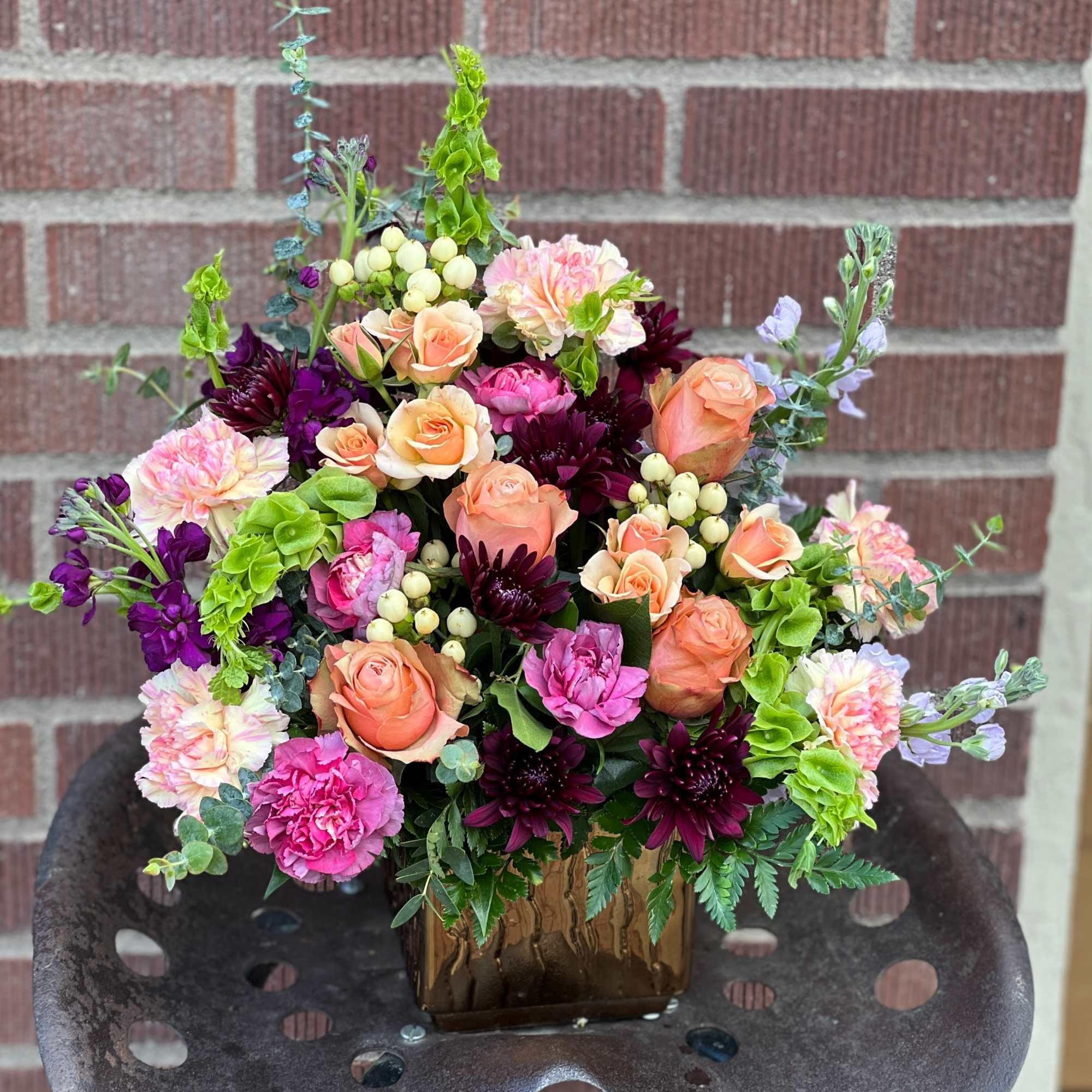 Mixed bouquet of peach roses, pink carnations, and dark purple blooms in a square vase