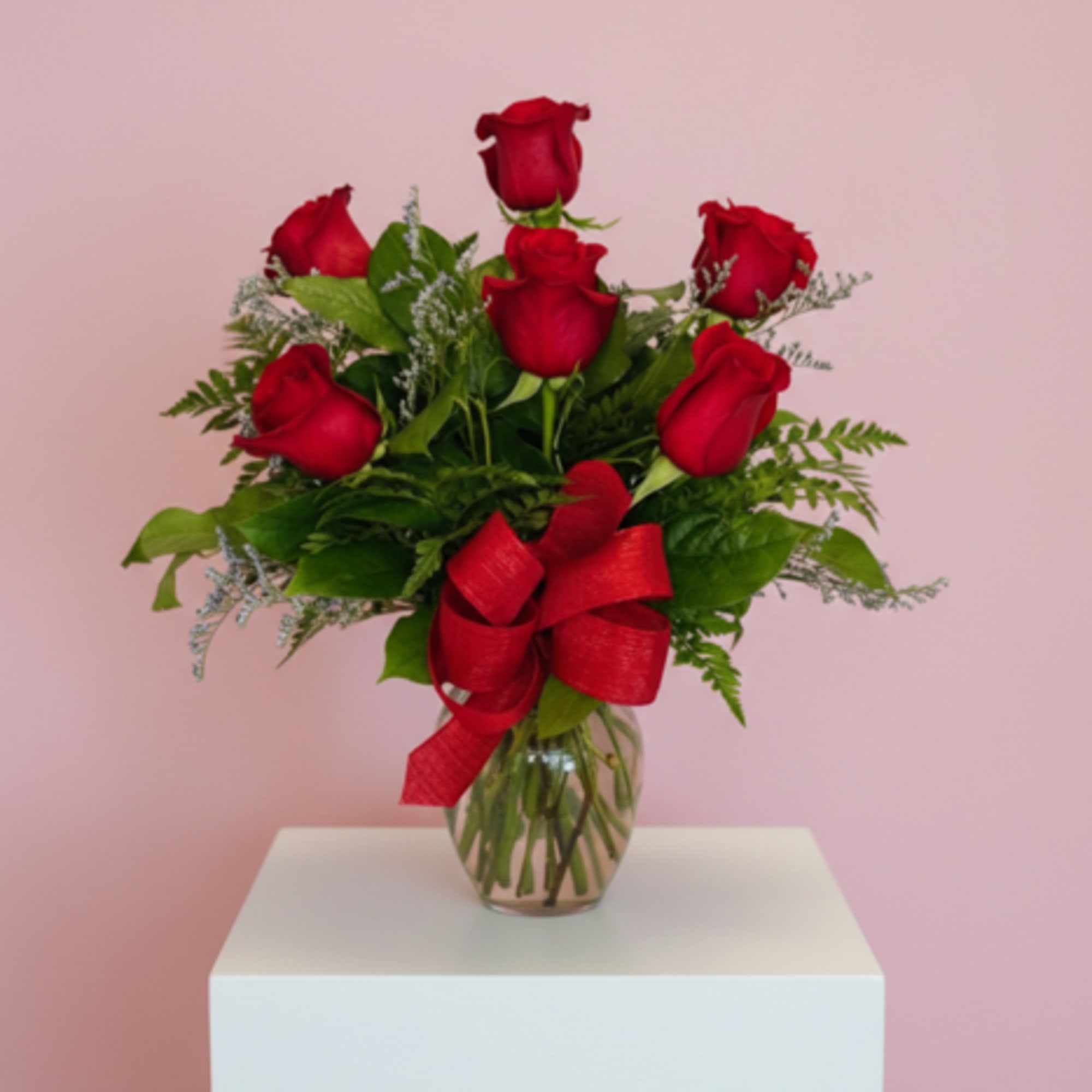 Red rose arrangement in a clear glass vase with a red bow on a white pedestal and pink background