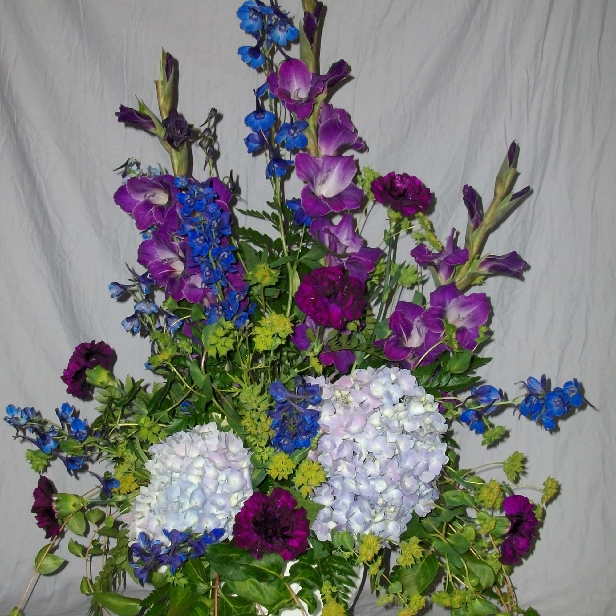 Tall floral arrangement with purple blooms, blue spikes, and pale hydrangeas in a white vase