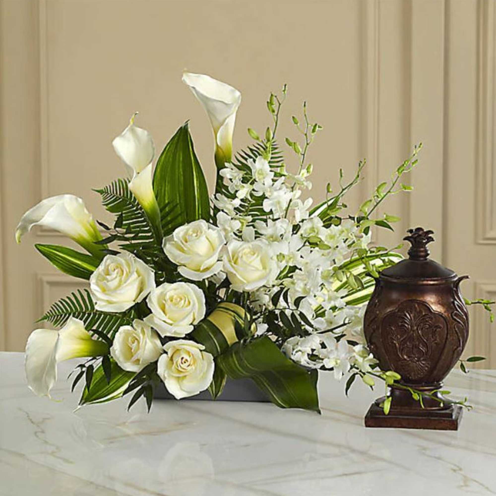 White roses and calla lilies arranged with greenery beside a dark urn.