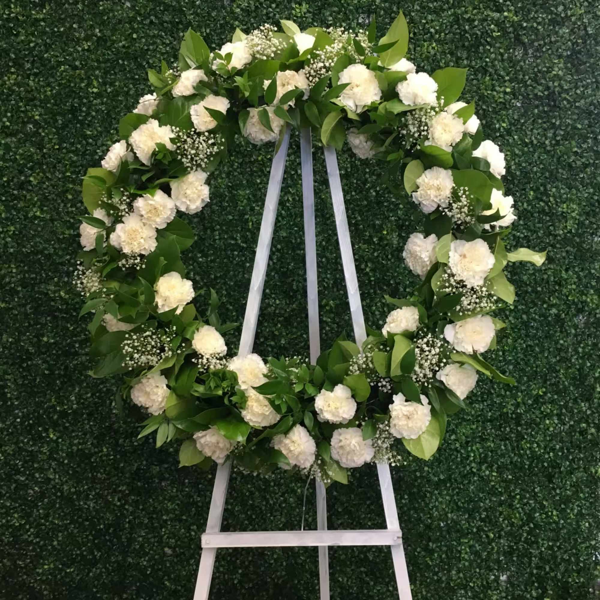 White floral wreath on a white easel against a green hedge wall