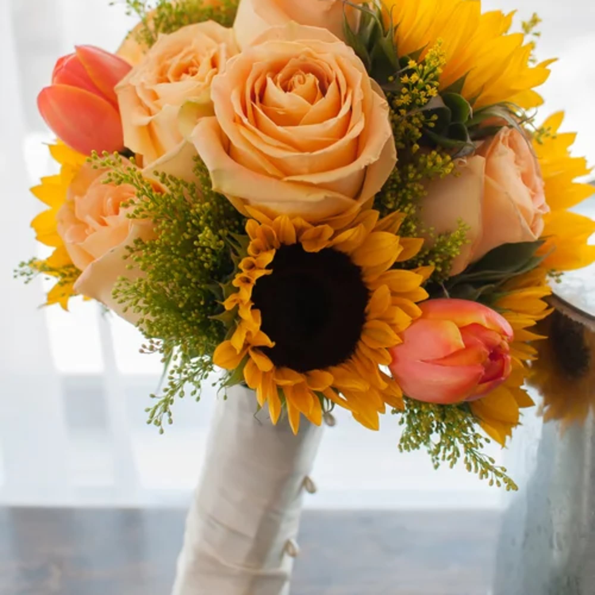 Bouquet of peach roses, sunflowers, and pink tulips wrapped in white ribbon