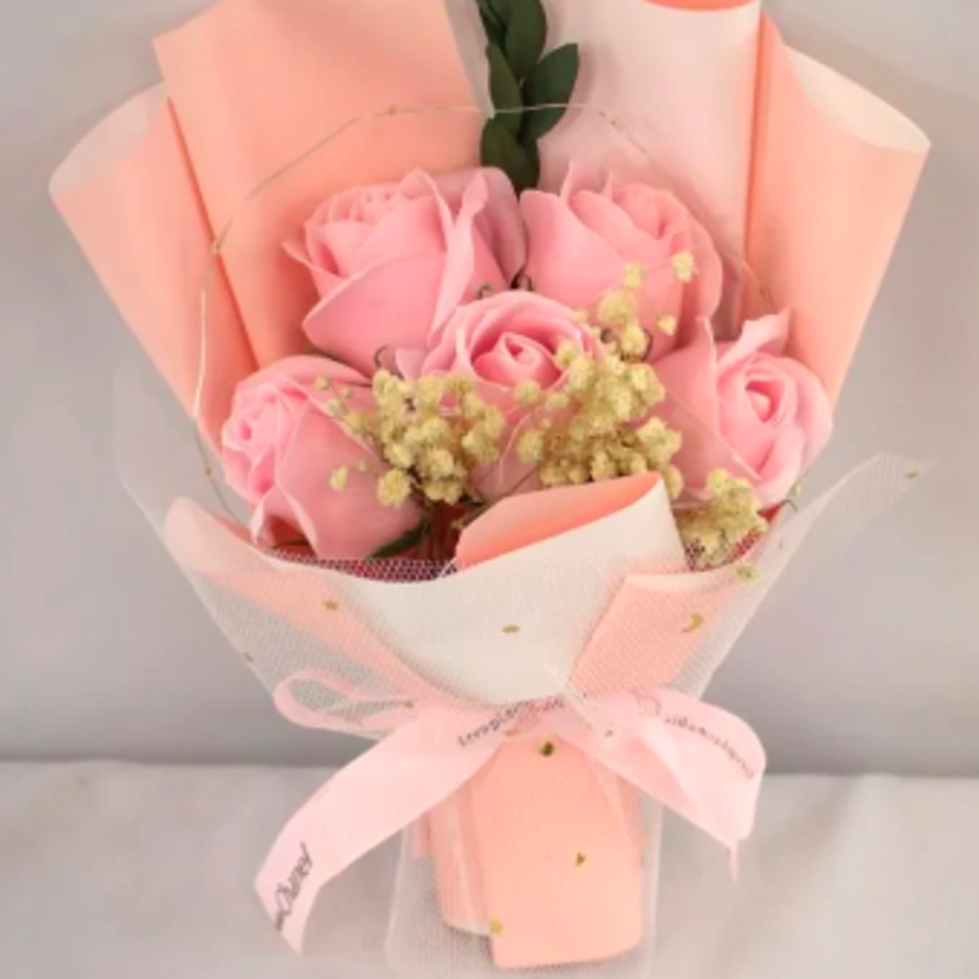 Small bouquet of pink roses with tiny cream filler flowers wrapped in peach paper and tulle with a pink bow.