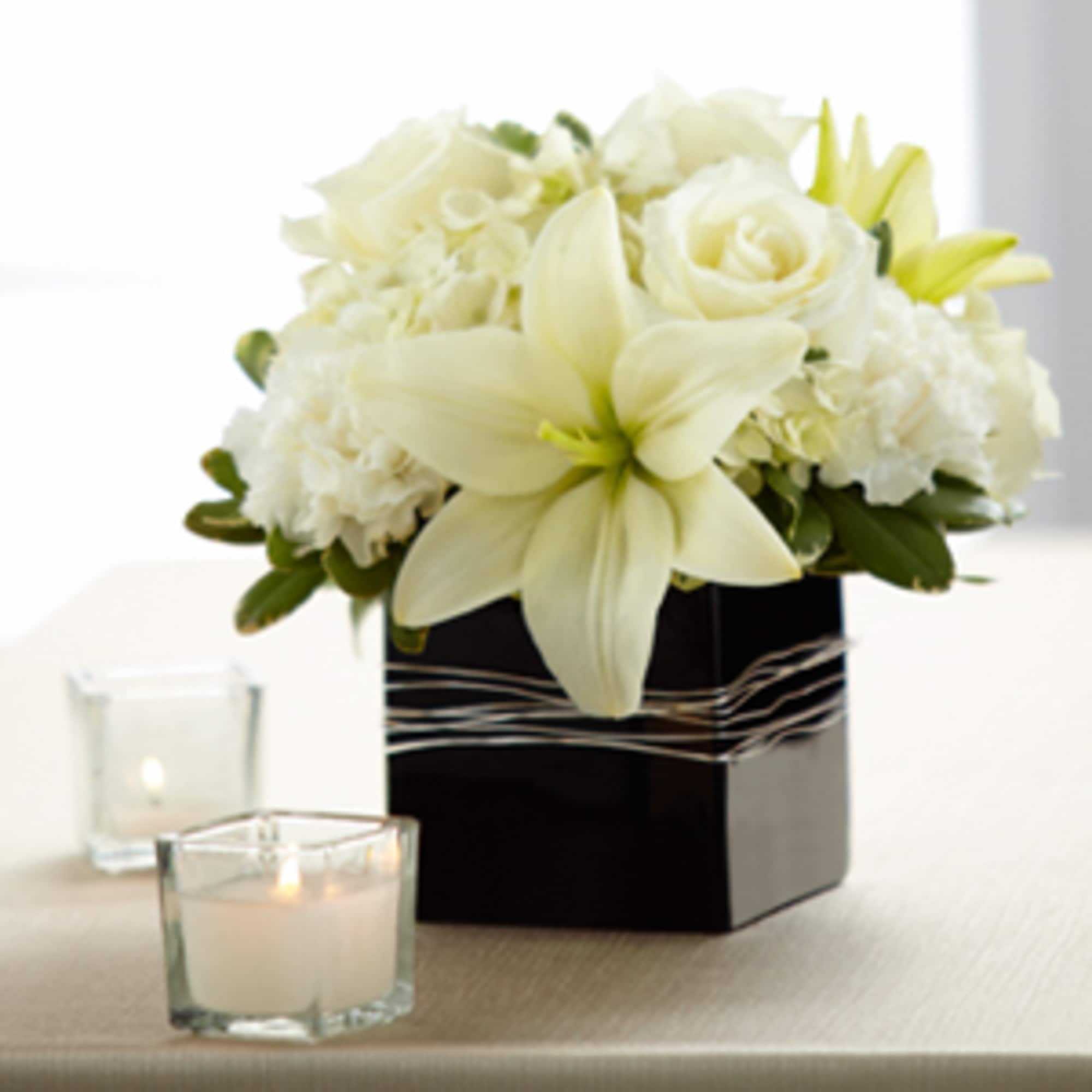 Low white lily and rose arrangement in a black square vase with votive candles