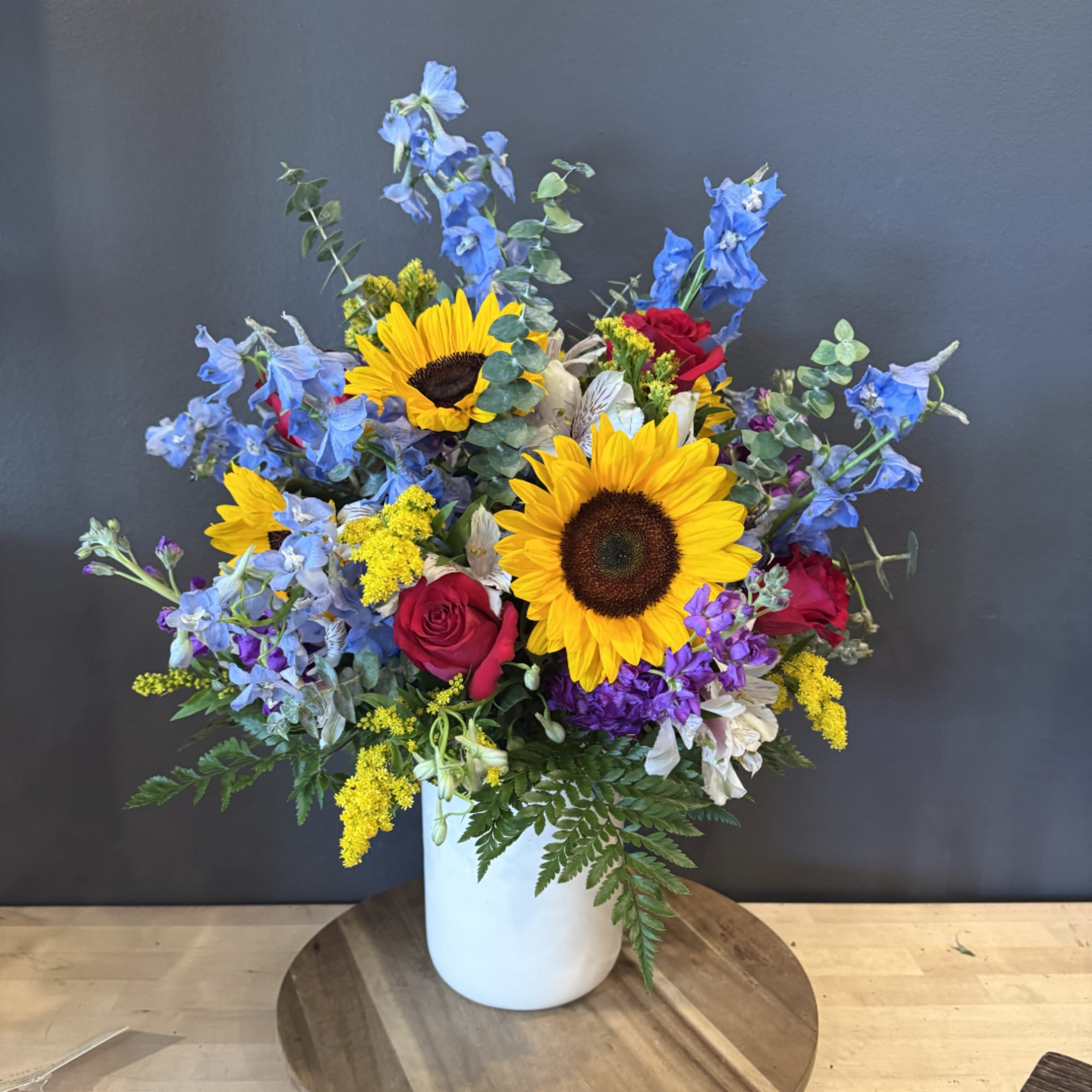 Mixed bouquet with sunflowers, blue delphinium, and red roses in a white vase