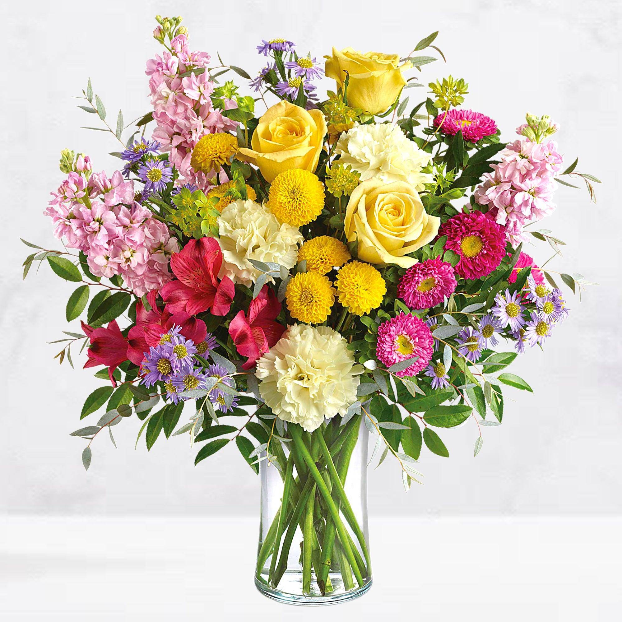 Mixed bouquet of yellow roses, pink blooms, and purple daisies in a glass vase