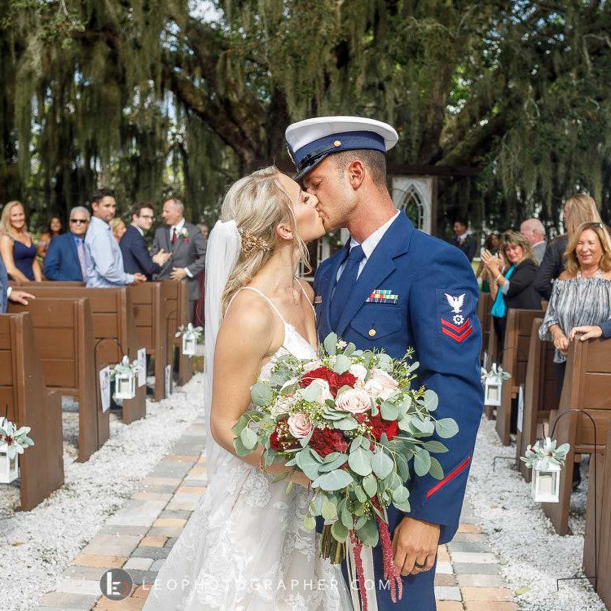 Bride and groom kissing in a wedding aisle with a bouquet of red, white, and blush flowers