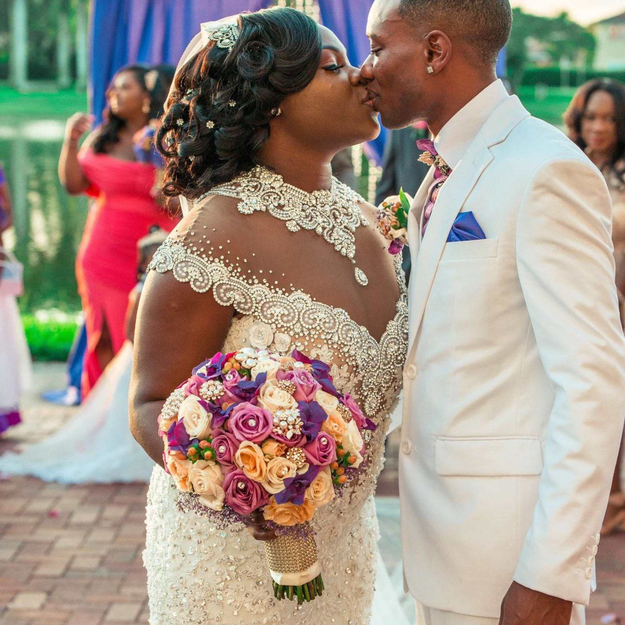 Bride holding a bouquet of pink, peach, and purple roses while kissing the groom