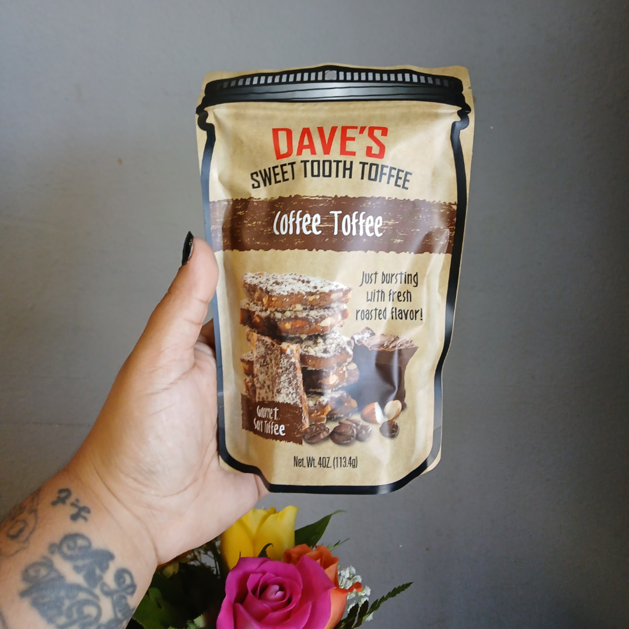 Hand holding a coffee toffee candy pouch above a small flower bouquet
