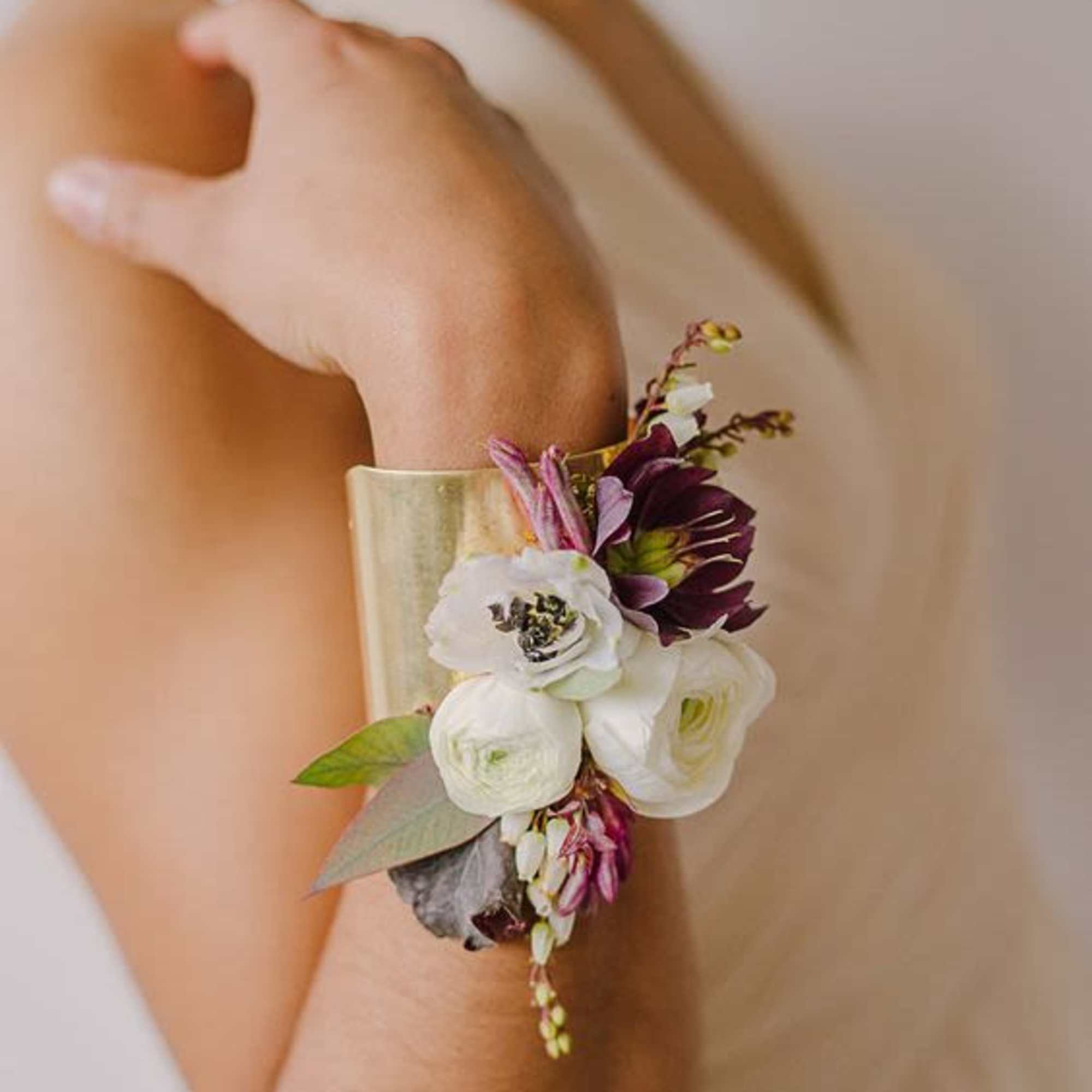 Gold cuff bracelet corsage with white and plum flowers worn on a person's forearm