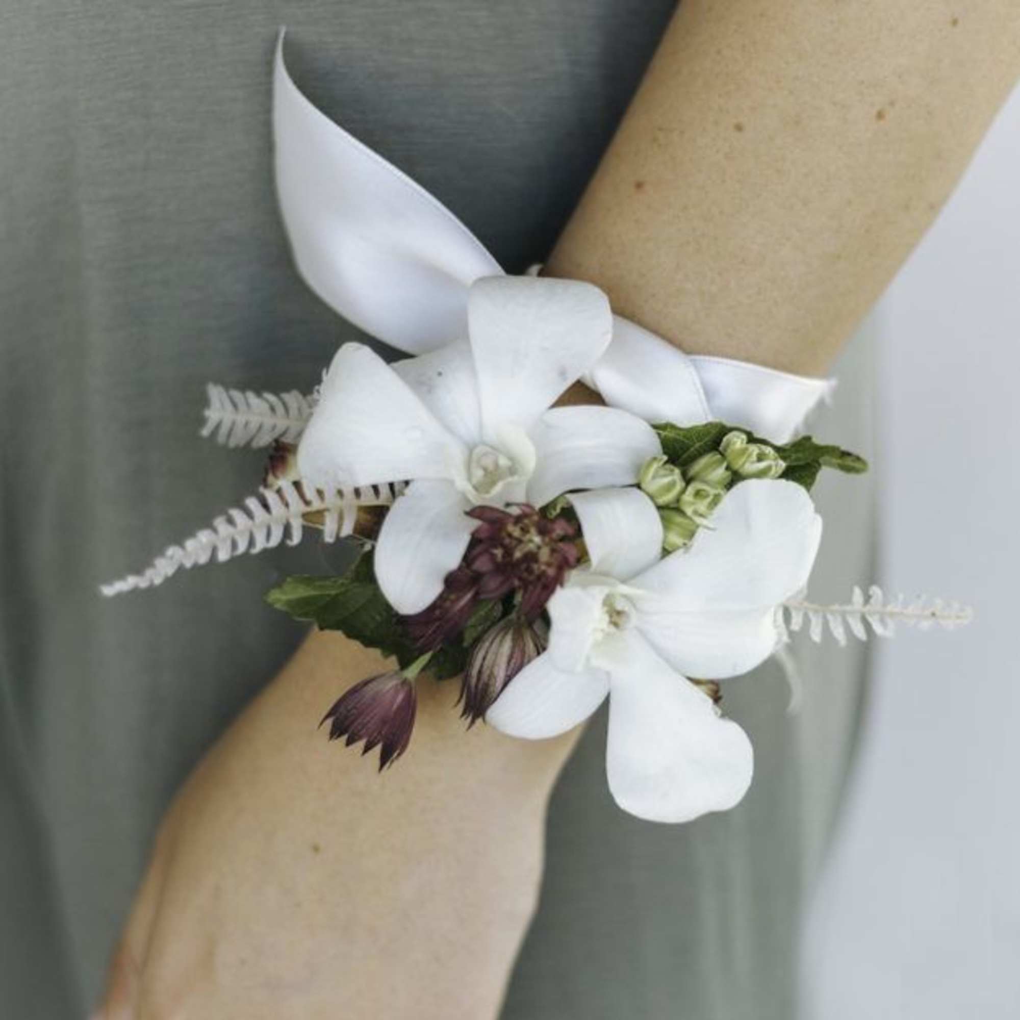 White orchid wrist corsage with purple buds on a white ribbon around a person’s wrist