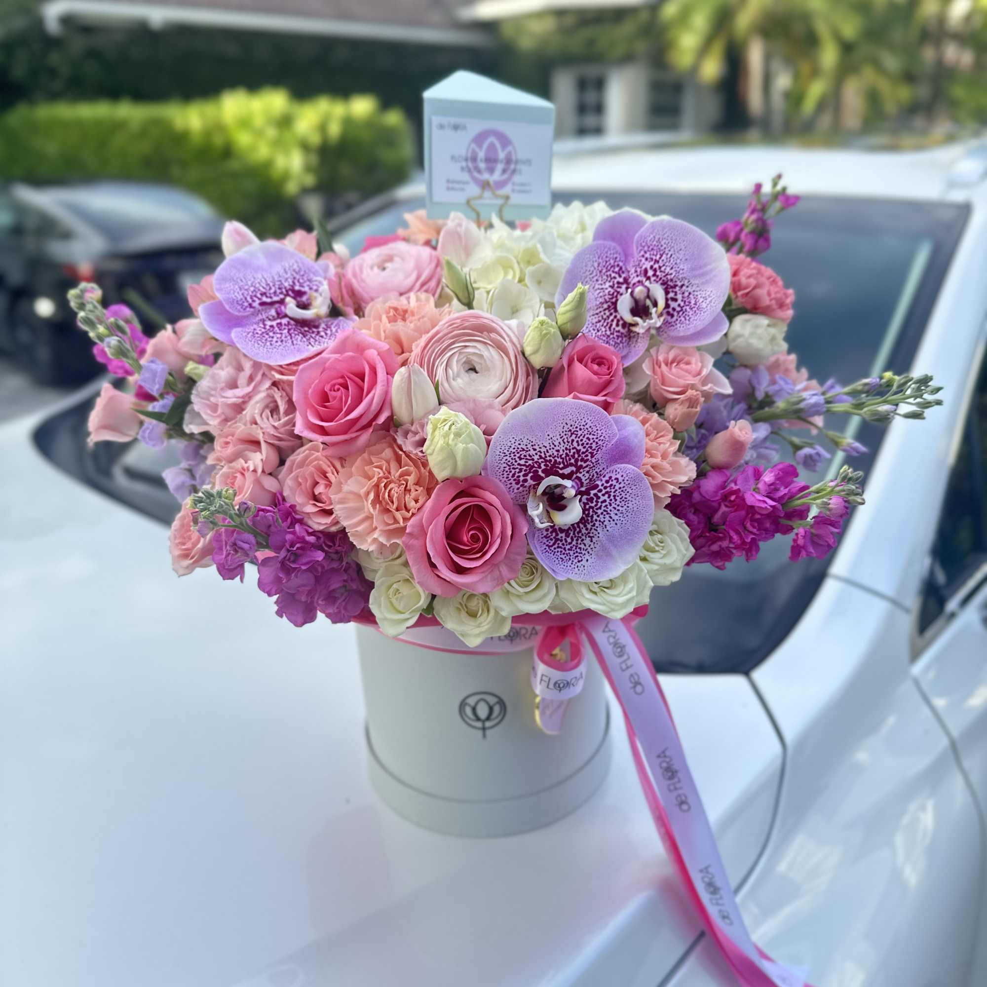 Round hatbox of pink and lavender roses, orchids, and mixed blooms with ribbon and card topper