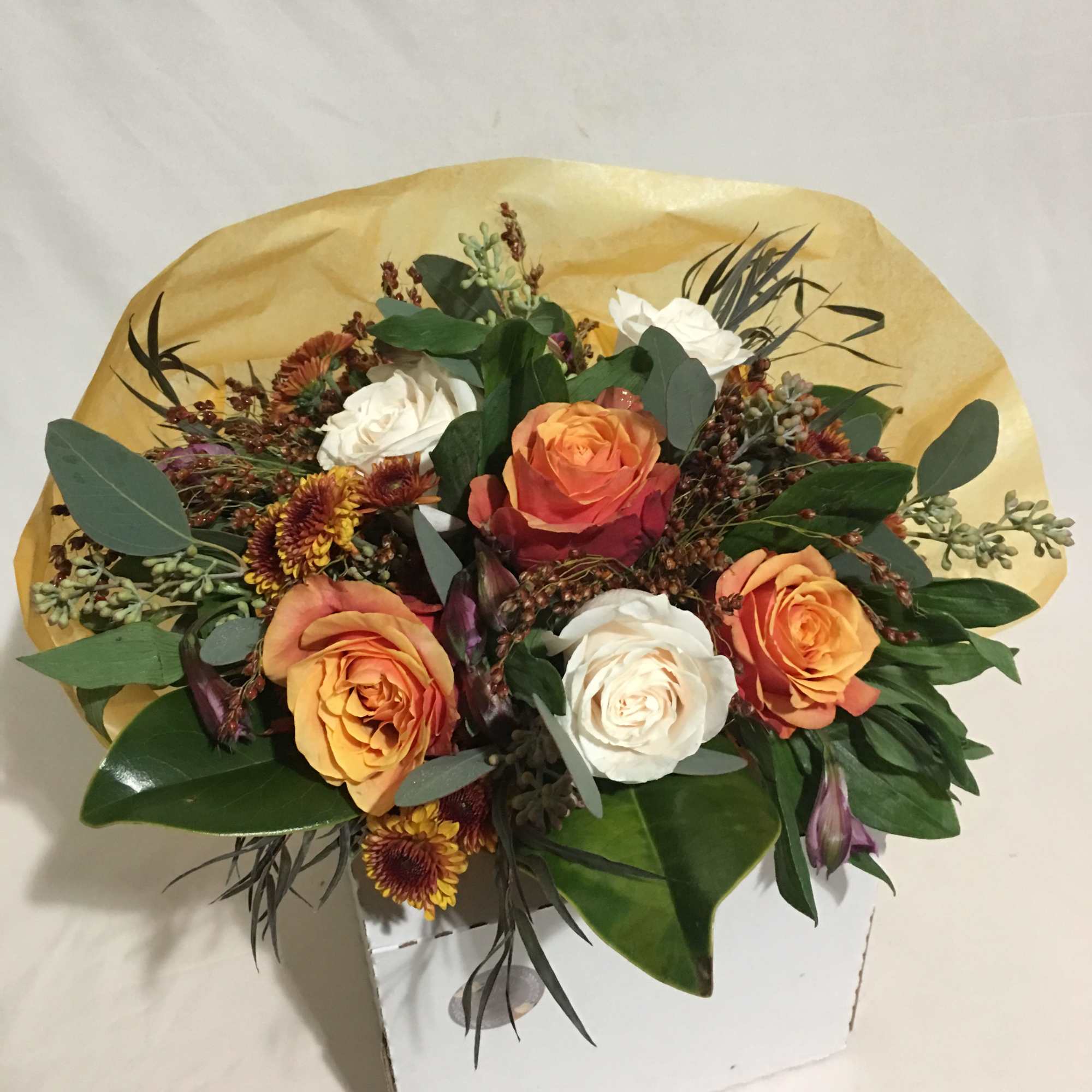 Hand-tied bouquet of orange and white roses with small rust flowers in yellow wrap on a white box