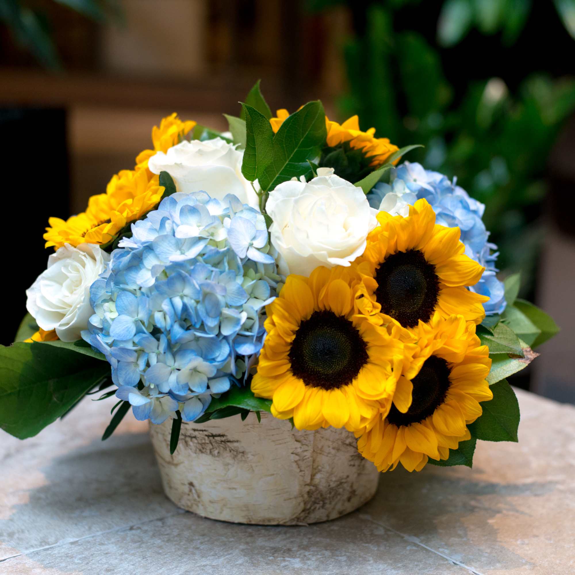Blue hydrangeas, white roses, and sunflowers in a low vase