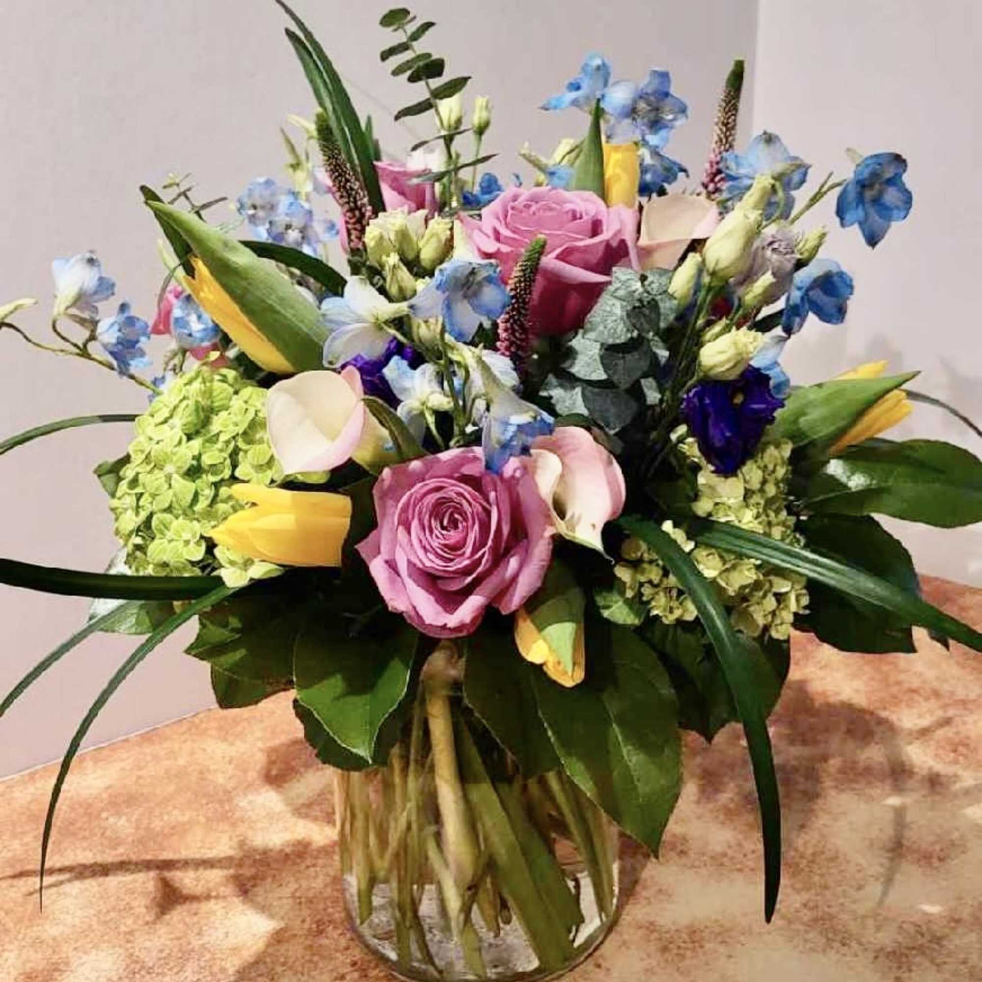 Mixed bouquet of pink roses, blue flowers, and yellow tulips in a glass vase