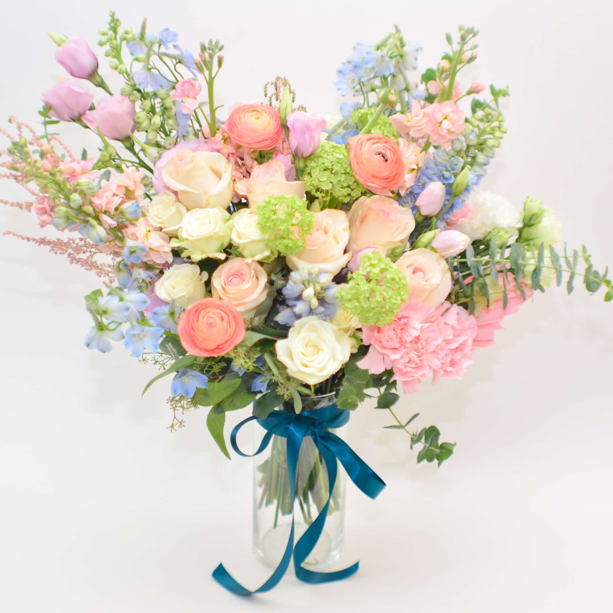 Pastel bouquet of roses, ranunculus, and blue flowers in a glass vase with a blue ribbon.