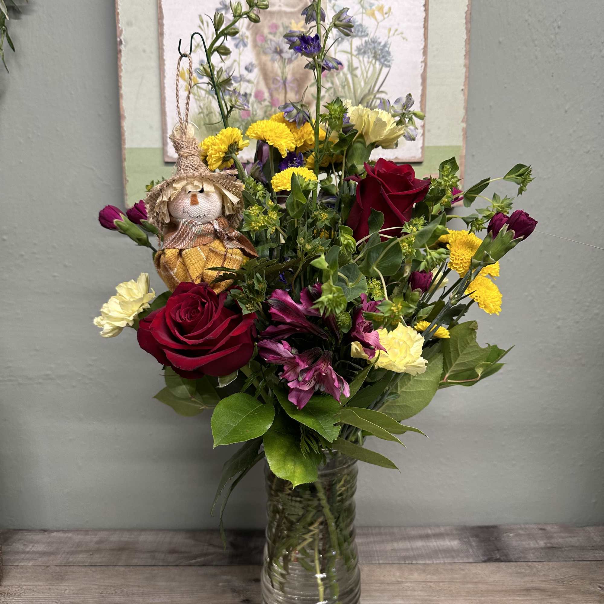Mixed bouquet of red roses, yellow blooms, and purple flowers in a glass vase with a small scarecrow pick