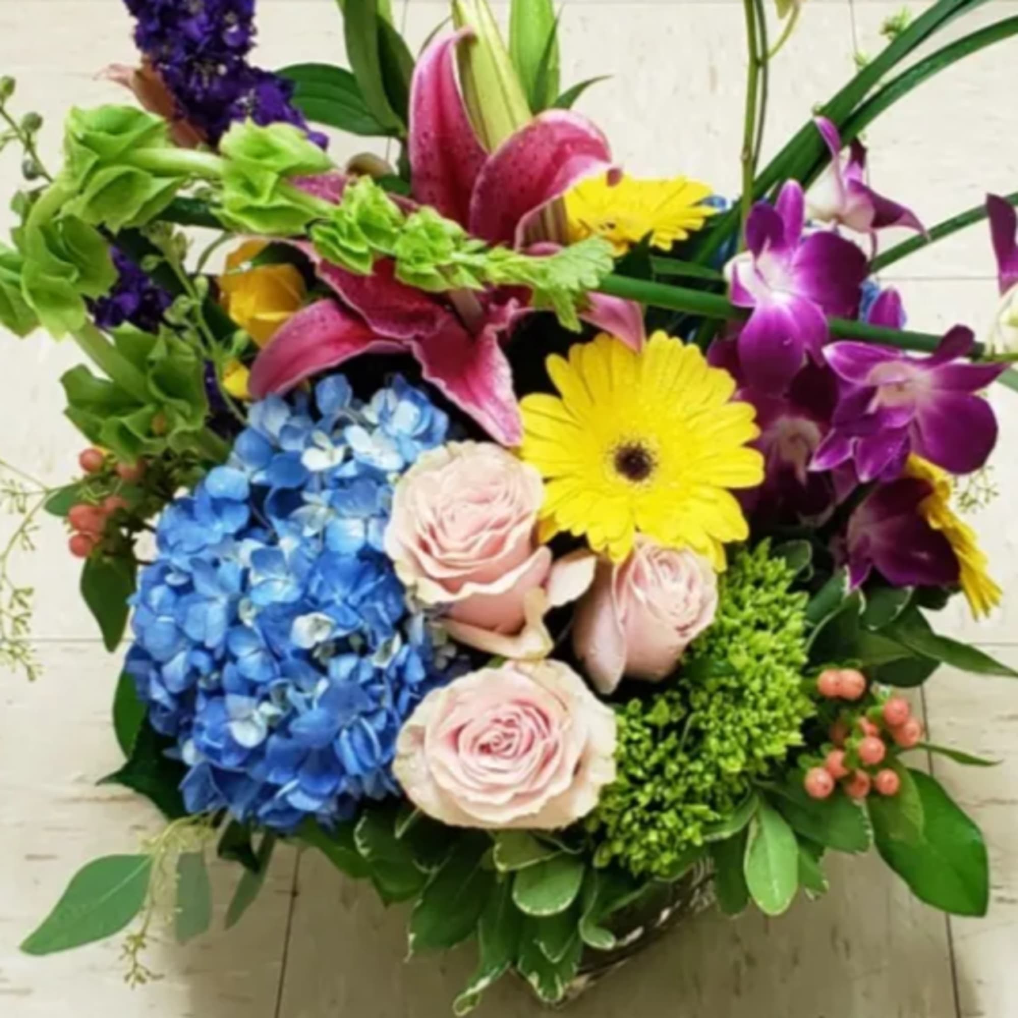 Colorful mixed arrangement with pink lilies, blue hydrangea, roses, yellow gerbera and orchids in a glass vase