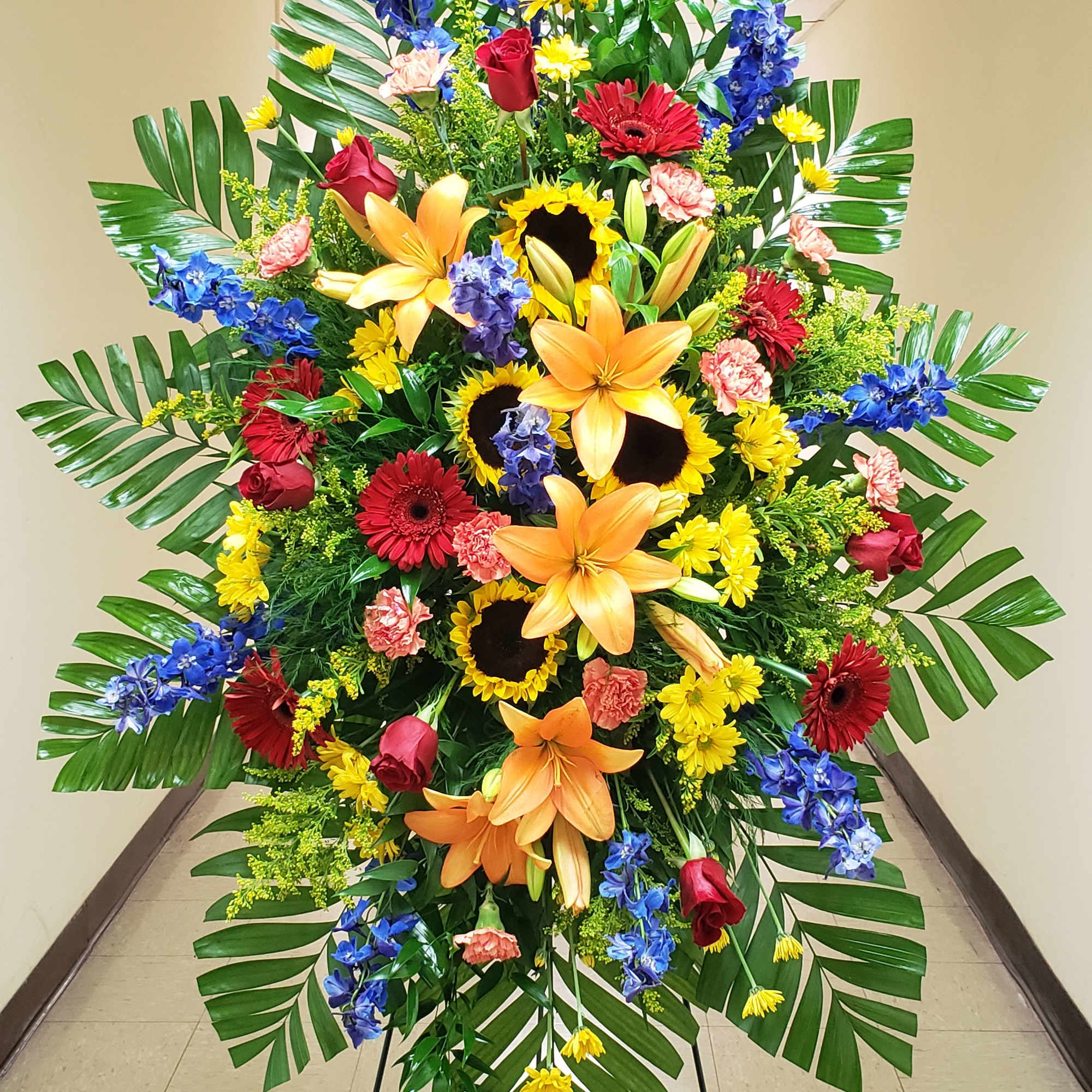 Tall standing spray with yellow lilies, sunflowers, red roses, and blue flowers on a metal easel.