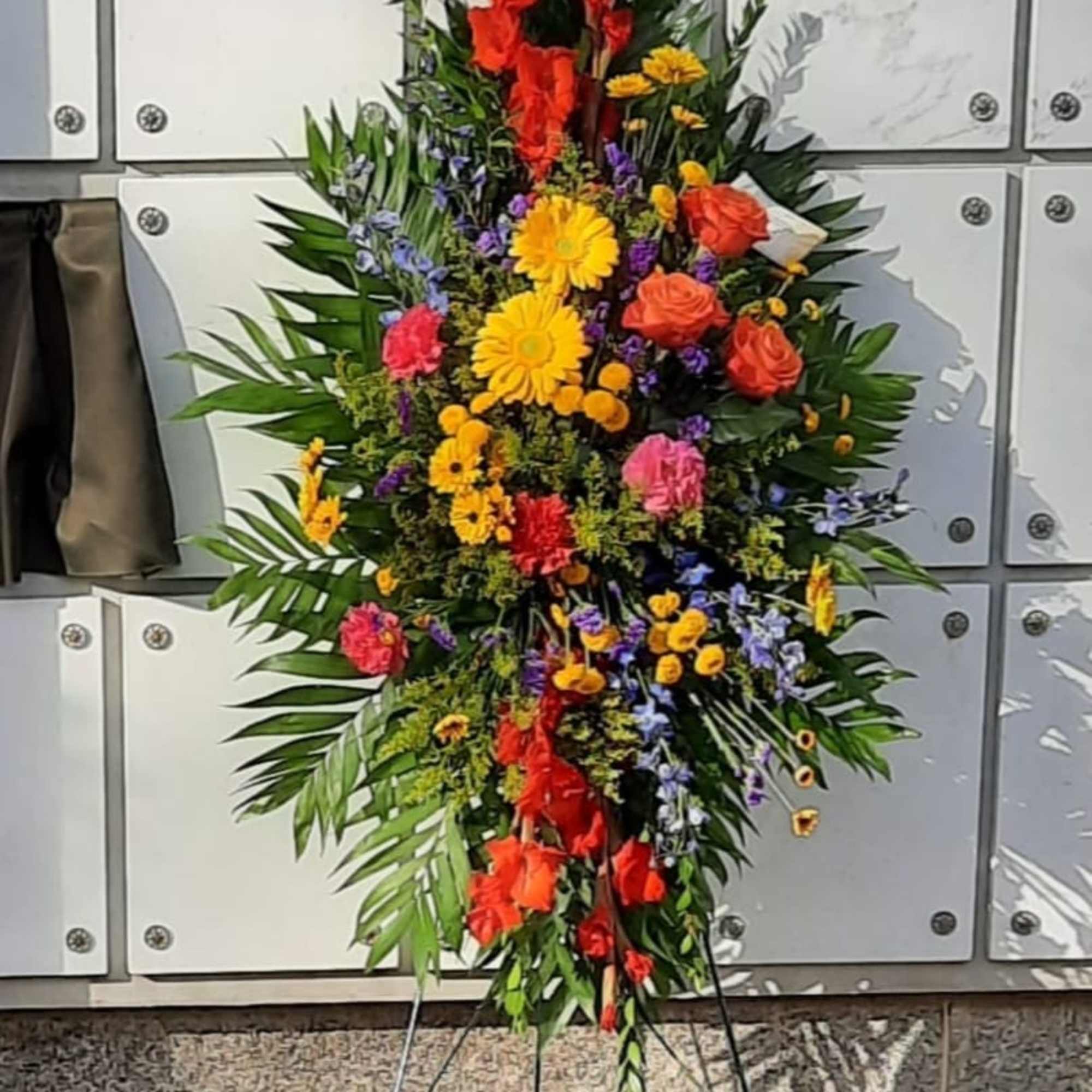 Tall standing spray of red, yellow, orange, and pink flowers on a metal easel against a white niche wall
