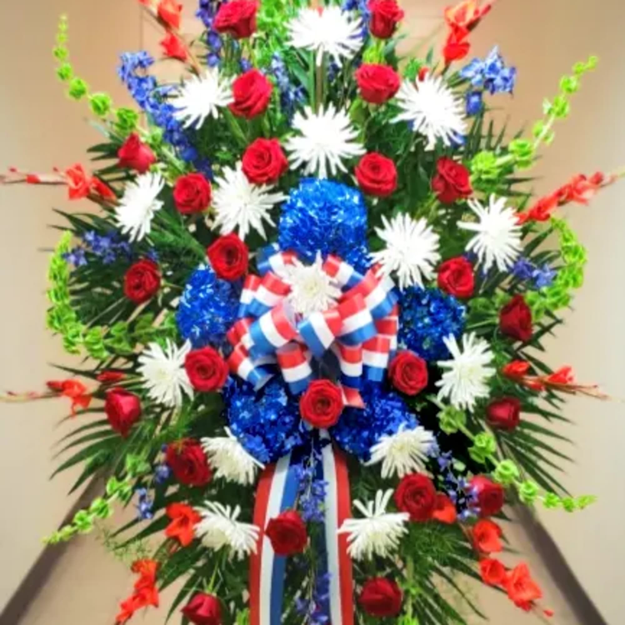 Tall patriotic standing spray with red roses, white mums, blue flowers, and a red, white, and blue ribbon bow