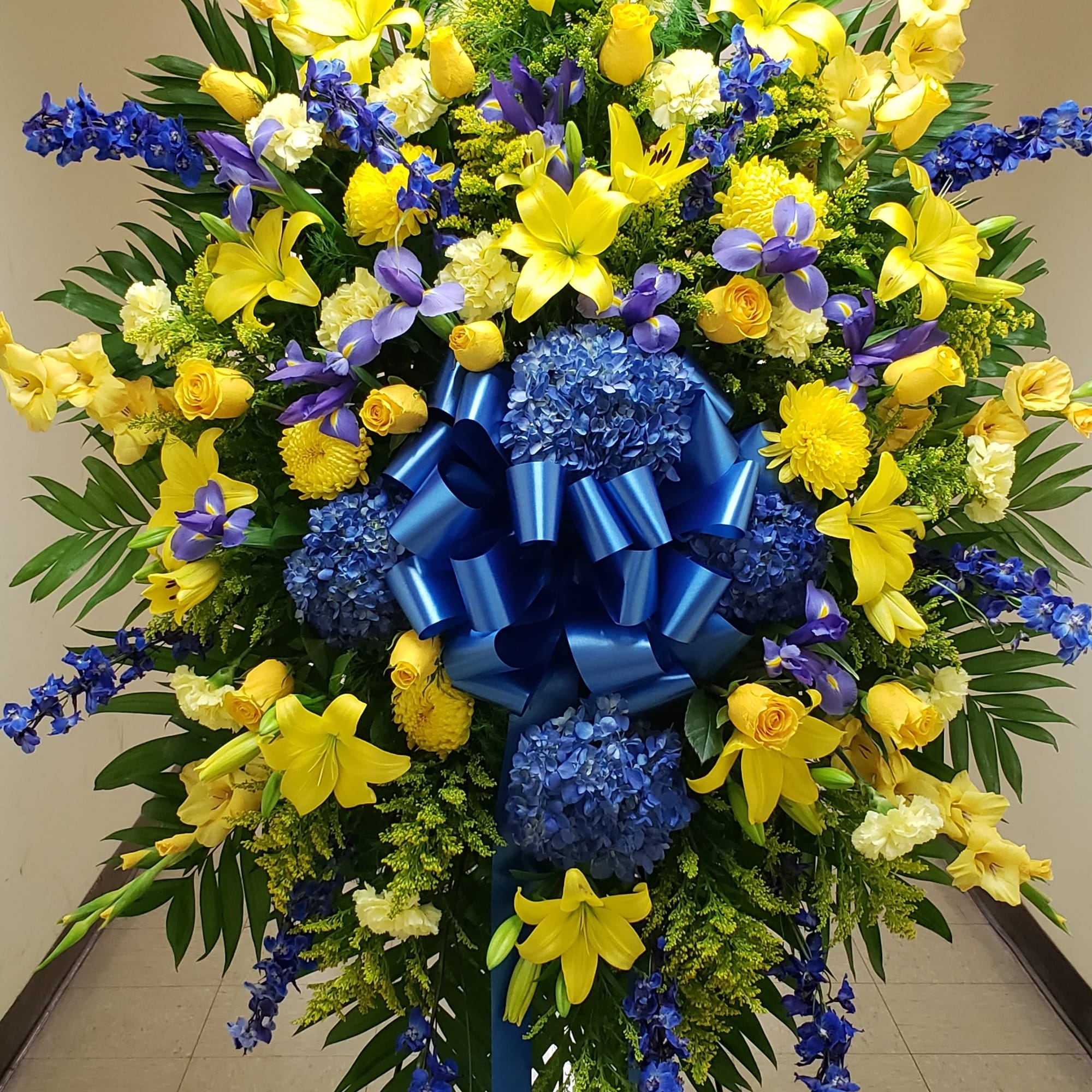Large standing floral spray with yellow and blue flowers and a blue ribbon bow