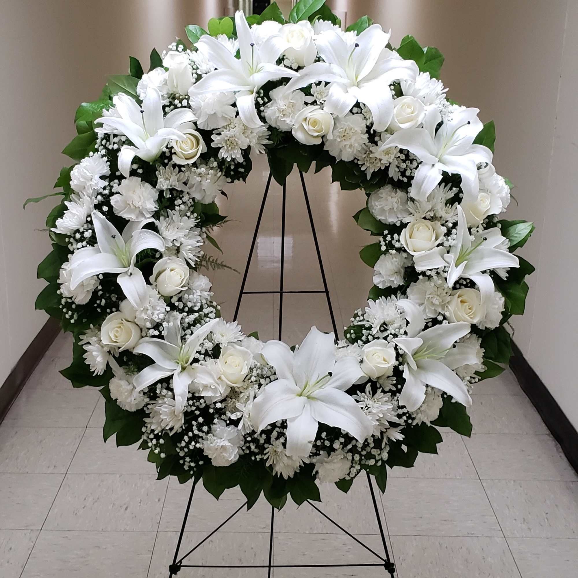 Large white floral wreath with lilies and roses displayed on a metal stand in a hallway.