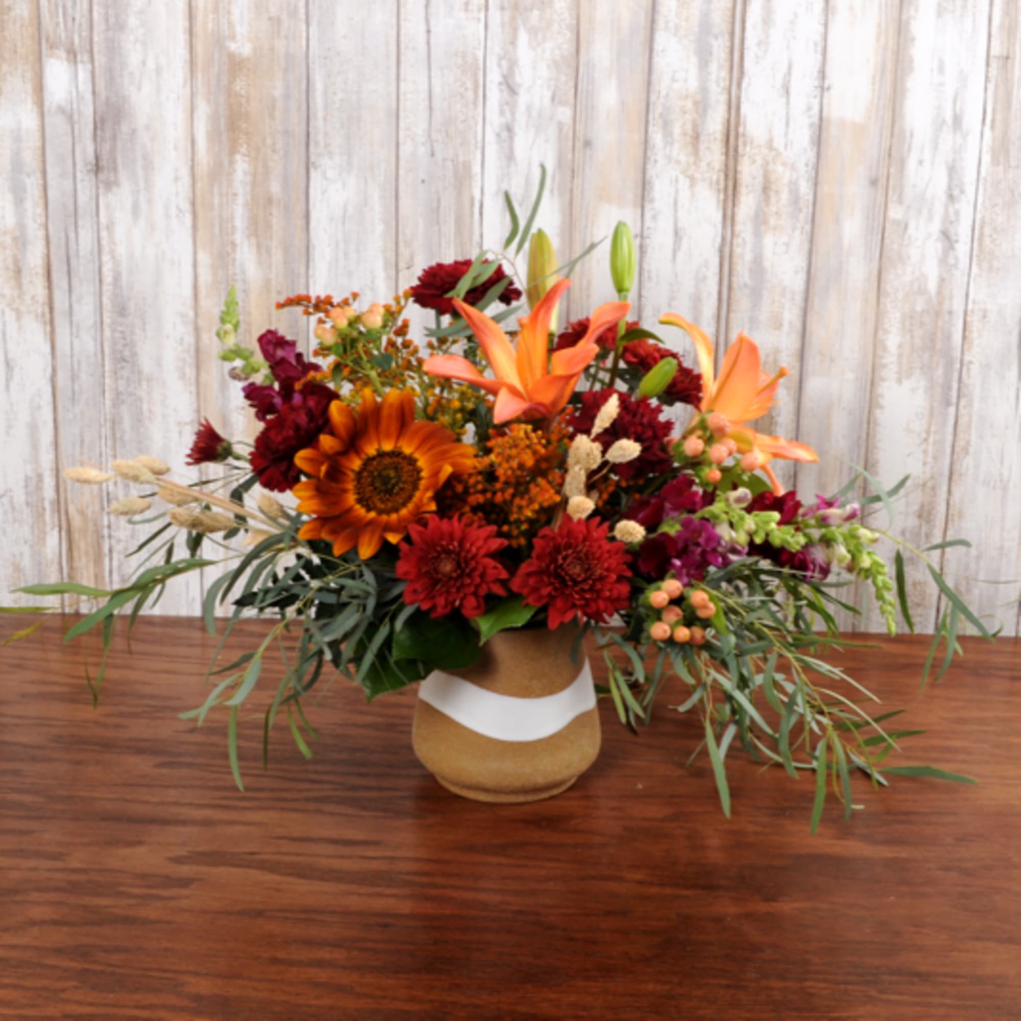 Low arrangement of orange lilies, sunflower, and red mums in a tan ceramic vase