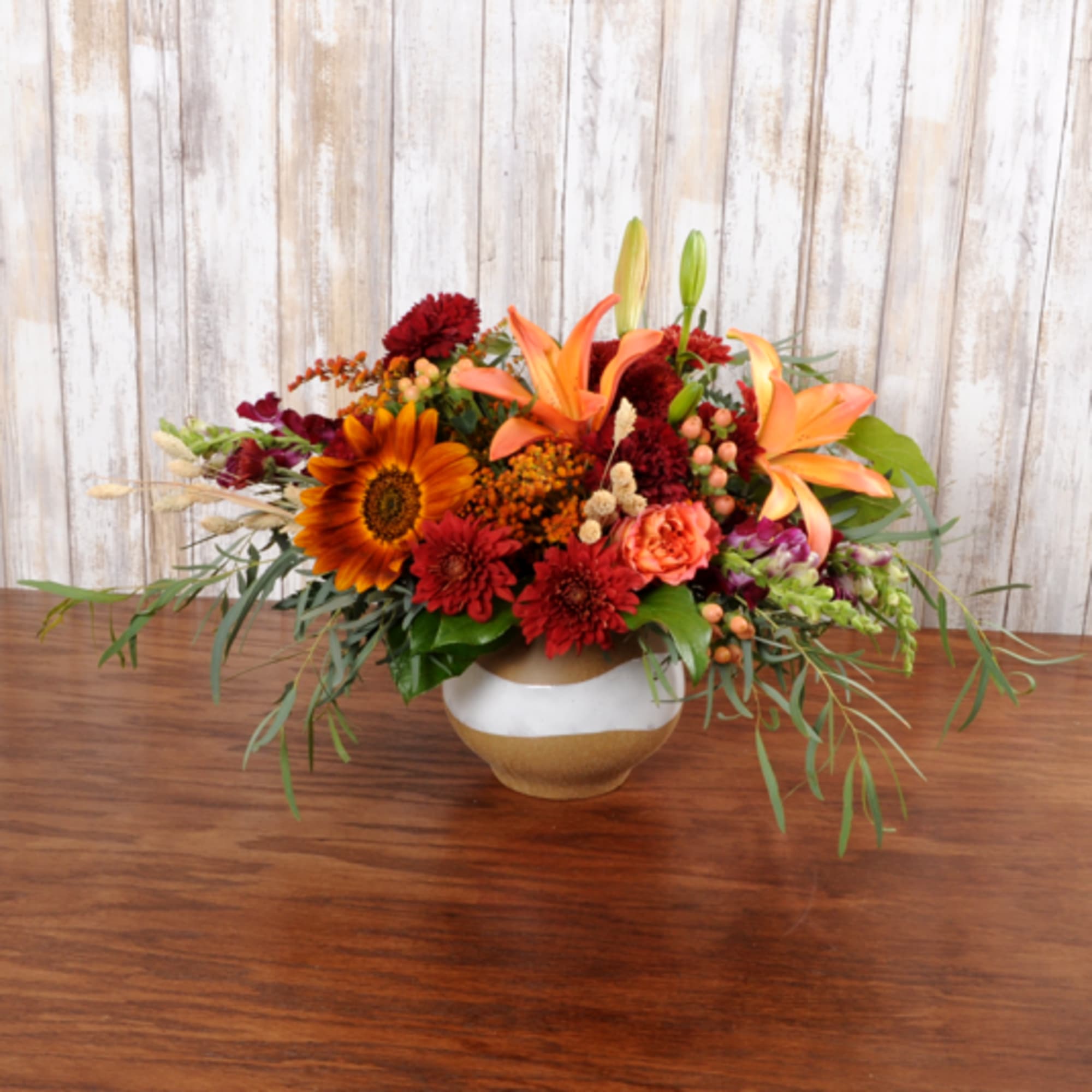 Low orange and red flower arrangement with lilies and a sunflower in a round ceramic bowl