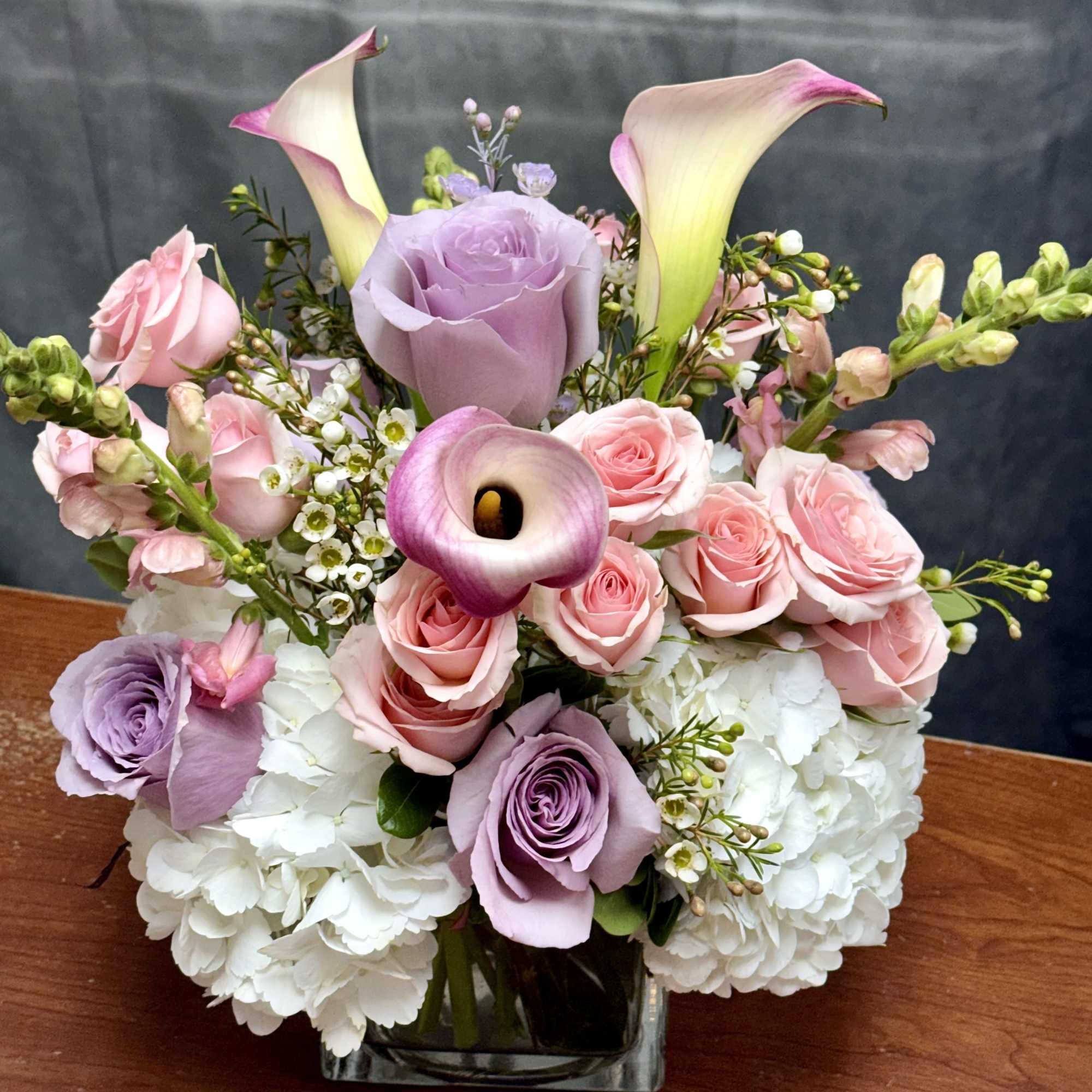 Pink and lavender roses with white hydrangeas and calla lilies in a glass vase.