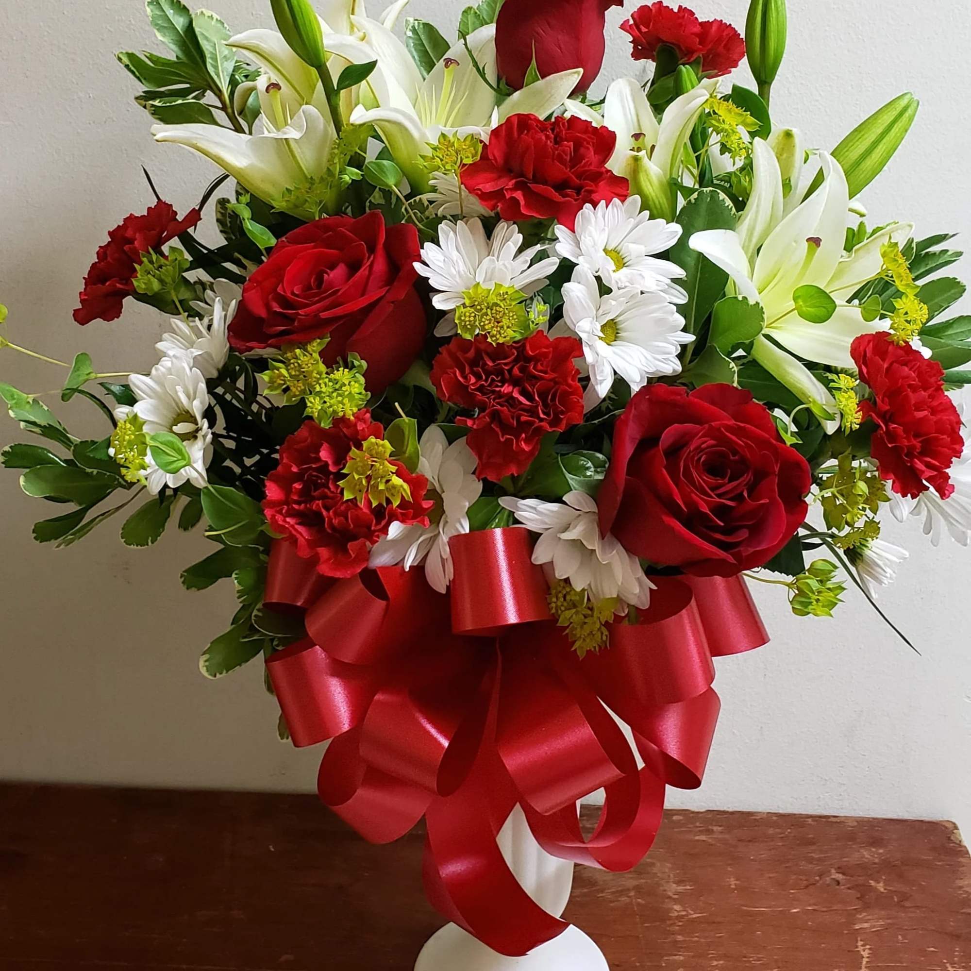 Red roses and white lilies arranged in a white vase with a red ribbon.