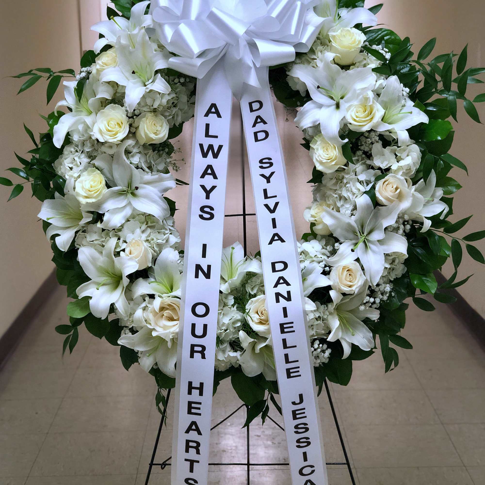White funeral wreath with lilies, roses, and a large bow on a stand