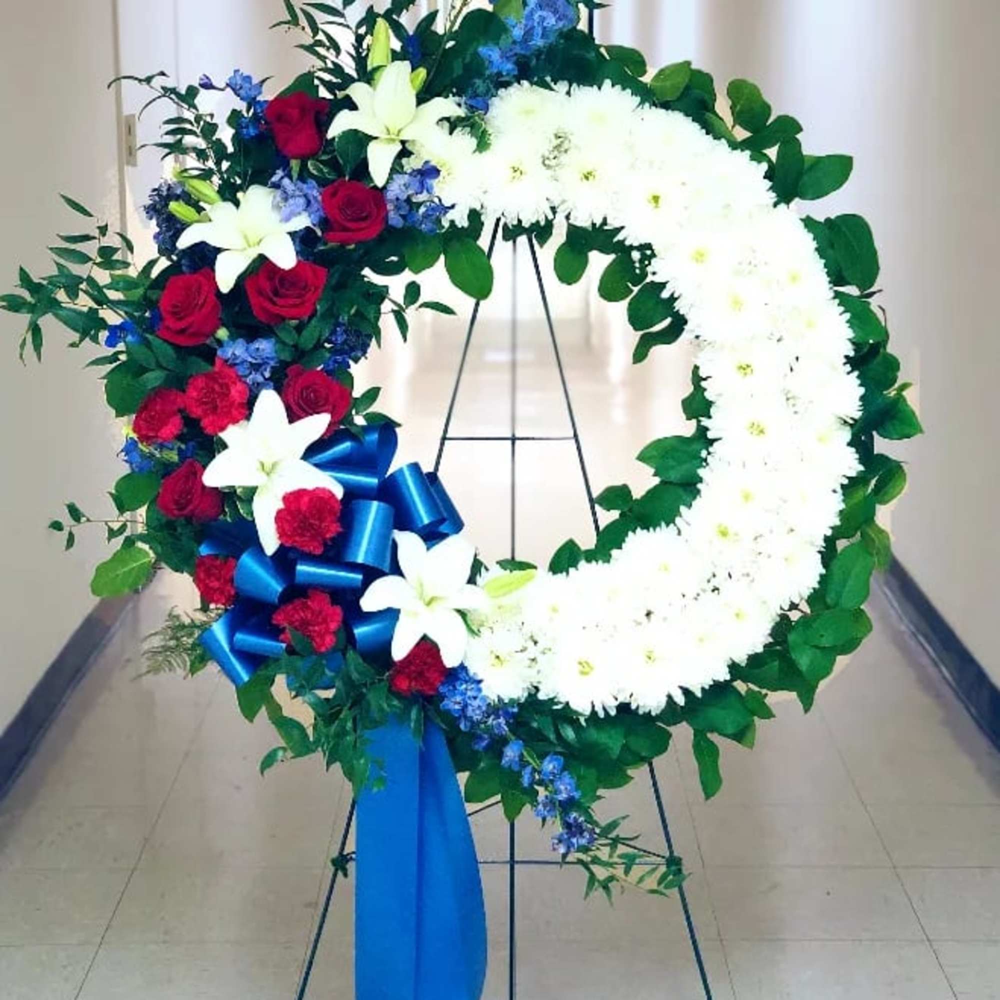 Large floral wreath with white chrysanthemums, red roses, blue flowers, and a blue ribbon