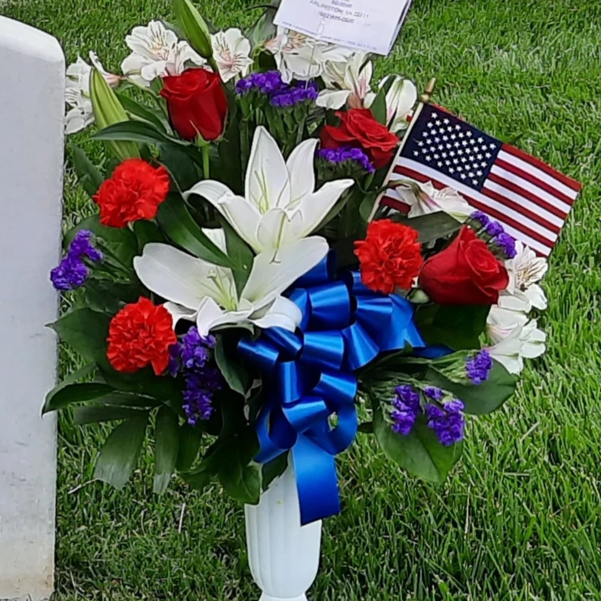 Red roses, white lilies, and purple flowers in a white vase with a blue ribbon and U.S. flag.