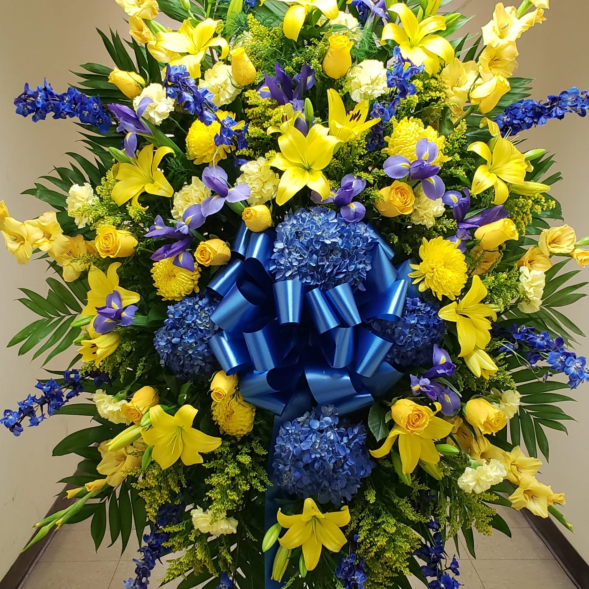 Large standing floral spray with yellow and blue flowers and a blue ribbon bow