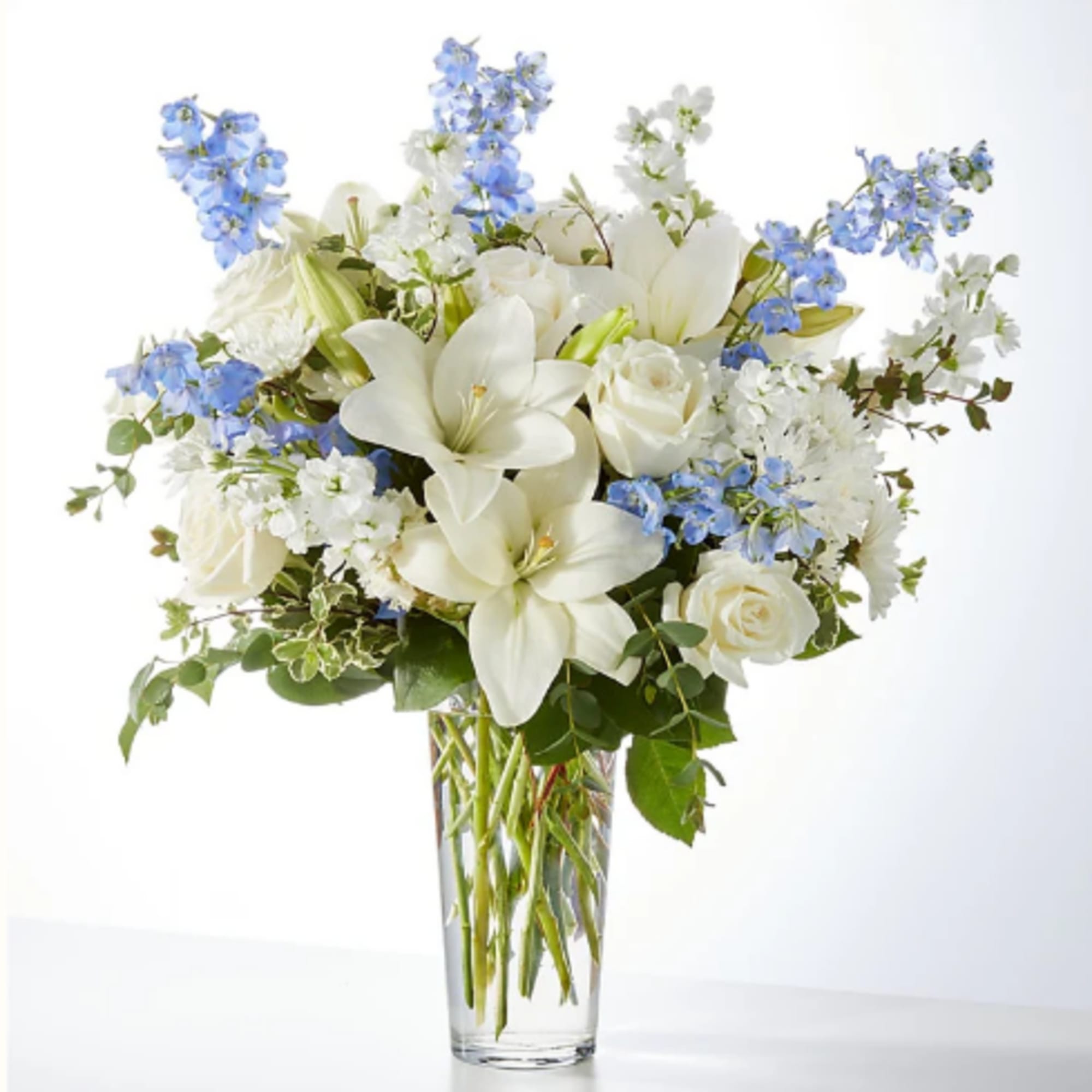 White lilies and roses with blue flowers in a clear glass vase