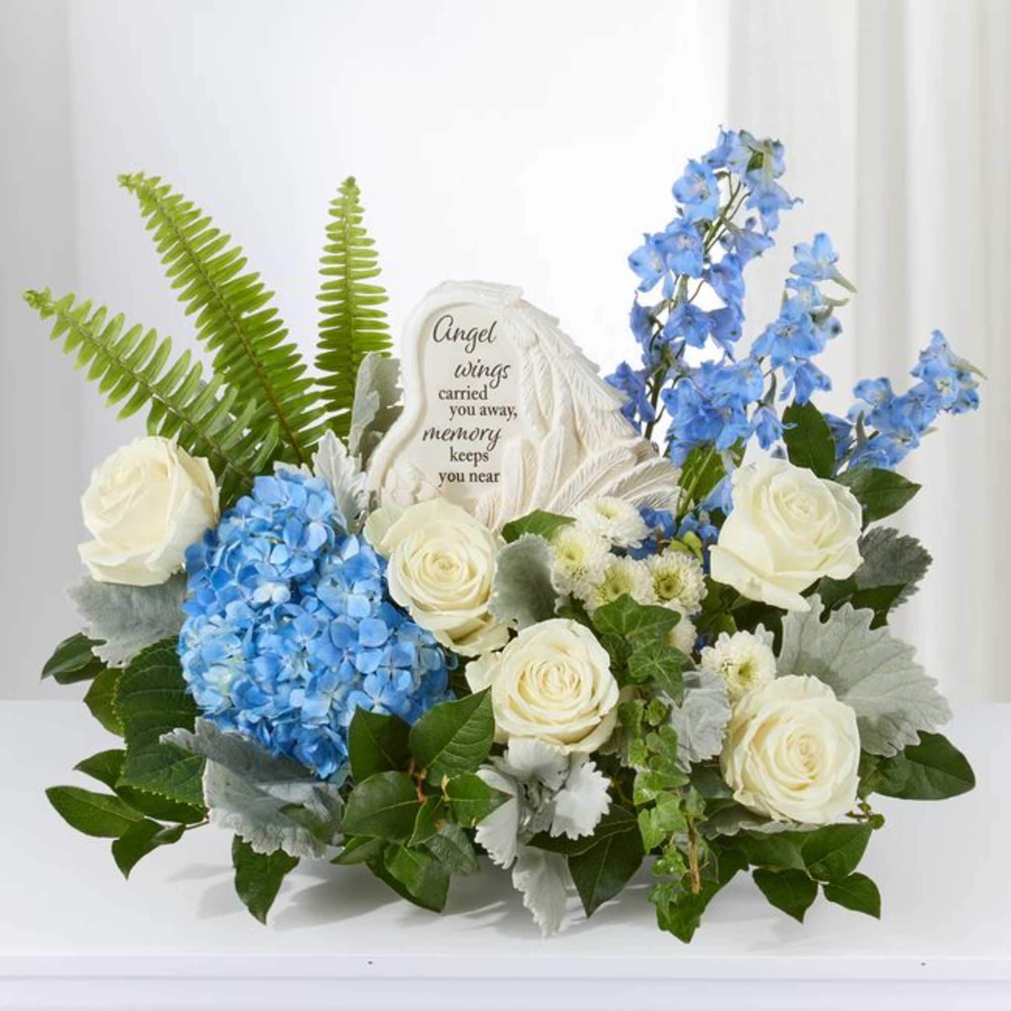 Sympathy arrangement with white roses, blue flowers, and an angel wing memorial plaque