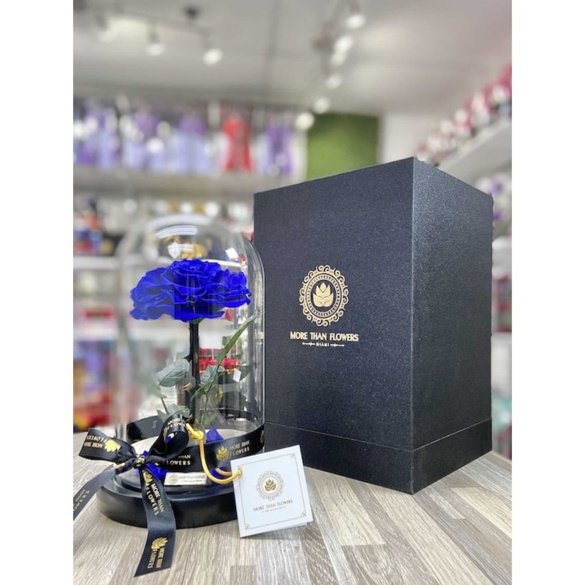 Single blue rose under a glass dome on a black base beside a tall black gift box.
