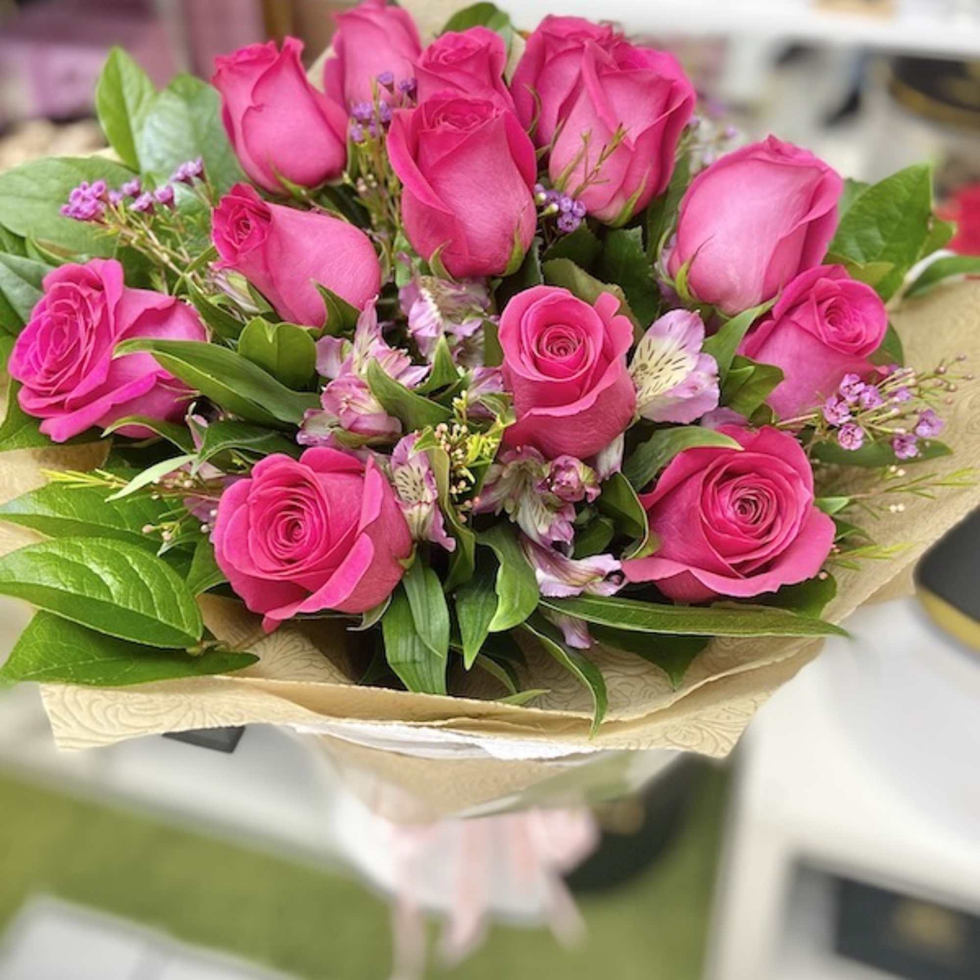 Hand-tied bouquet of bright pink roses with light pink accent flowers wrapped in patterned kraft paper.