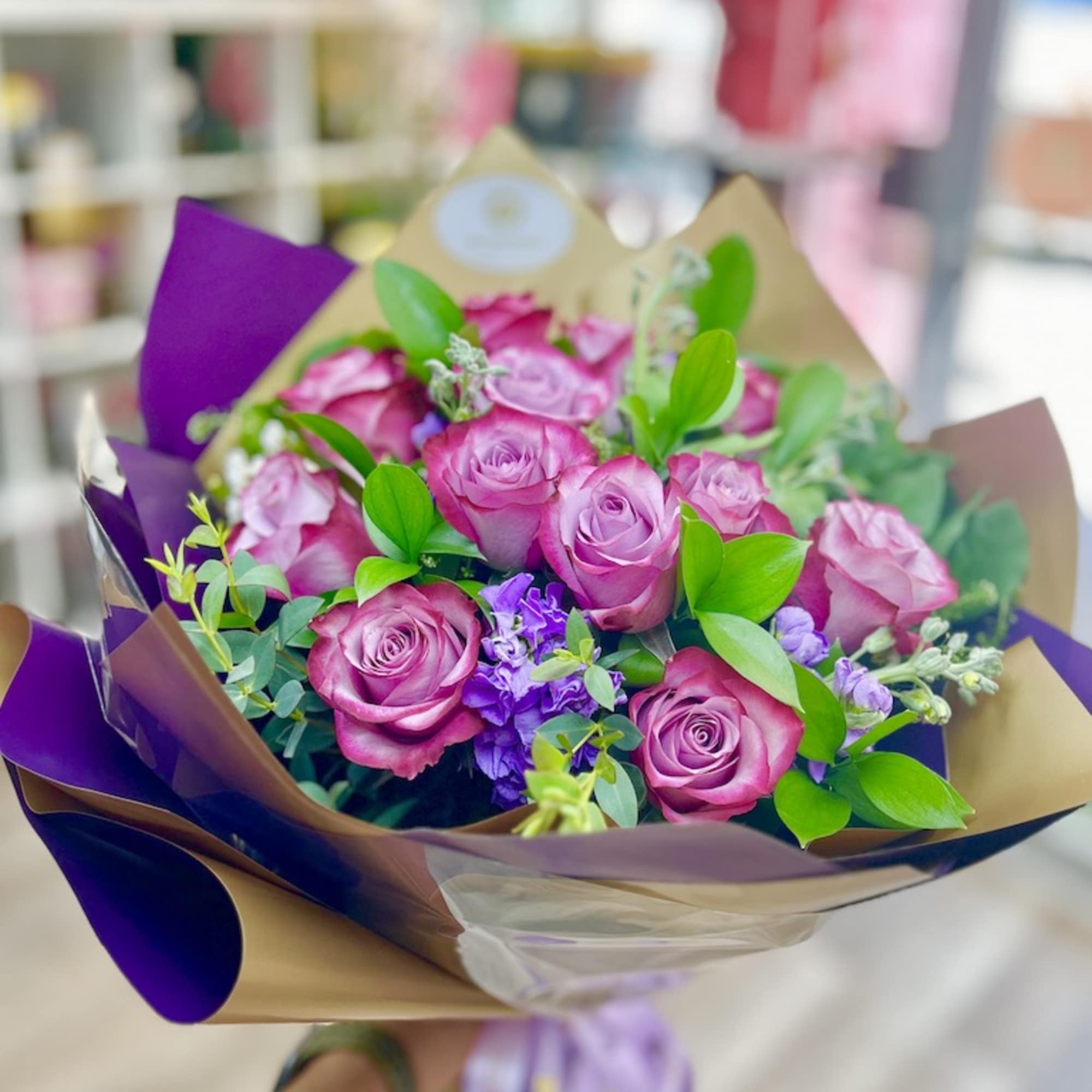 Handheld bouquet of pink and purple roses with small purple blooms wrapped in kraft and purple paper.